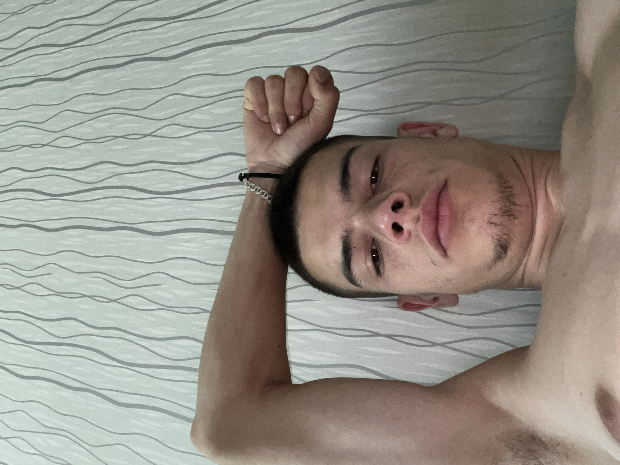 A young man, appearing to be in his late teens or early twenties, is captured in a selfie-style, close-up shot. He has short, dark hair and fair skin, with some visible stubble on his chin. His left arm is raised and bent, with his fist clenched and resting on his forehead. A silver chain bracelet is visible on his left wrist. The background consists of a wall with a light-colored, textured wallpaper featuring vertical, wavy grey lines. The lighting suggests it might be daytime, possibly indoors. There is no discernible information within the image to indicate a specific location in the city of Sopot, Bulgaria.