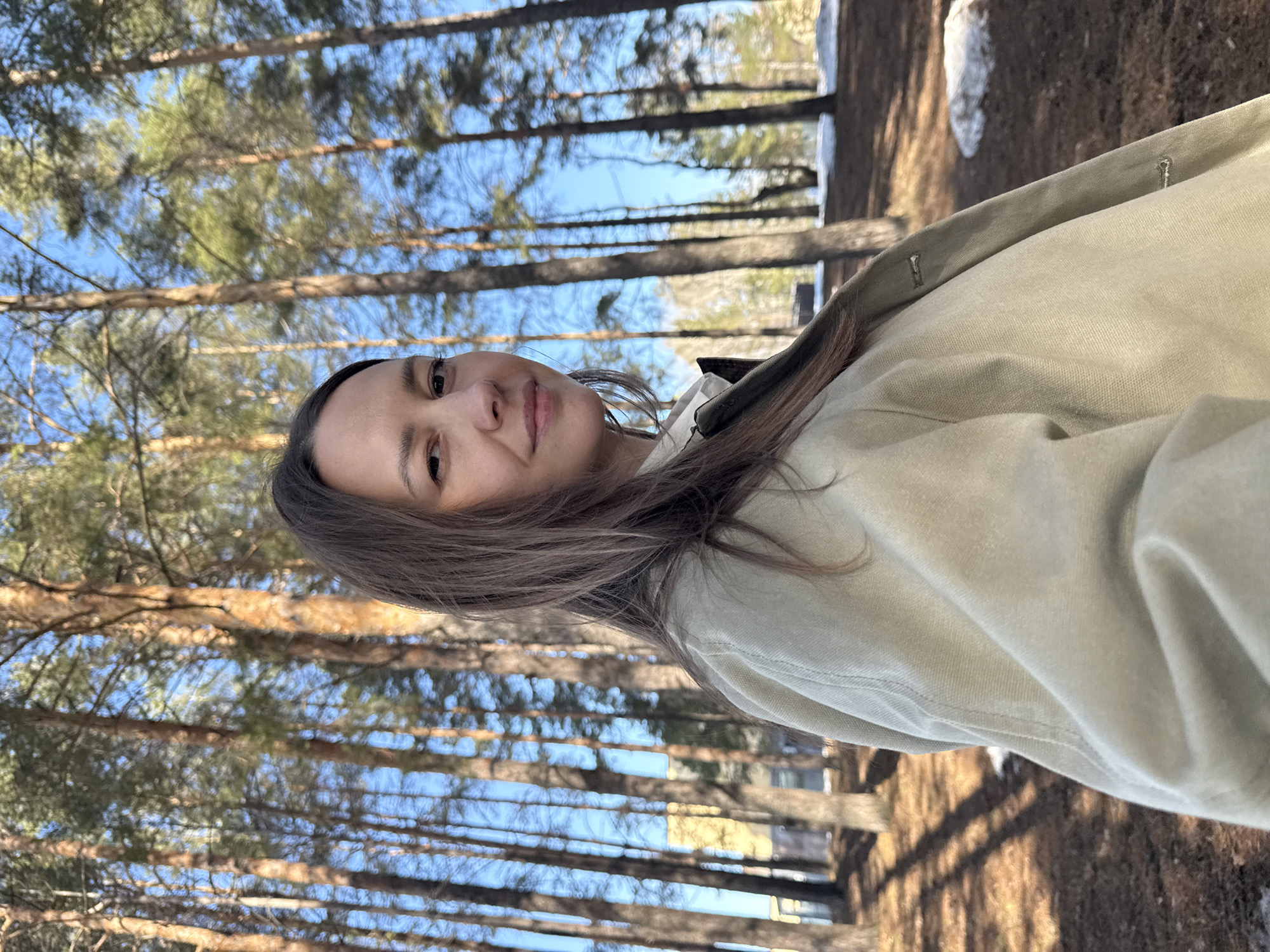 The image depicts a woman in an outdoor setting with a backdrop of tall pine trees. The location is identified as Chaikovsky, Russia, based on provided information.
**People:**
* A woman is the central subject of the image. She is facing the camera with a slight smile.
**Environment:**
* The scene is set in a forest or wooded area, characterized by tall pine trees with visible trunks and canopy against a blue sky.
* Patches of snow are visible on the ground, suggesting it is either late winter or early spring.
* The ground is covered with leaf litter.
**Objects:**
* The woman is wearing a light-colored jacket.
* The trees are tall and straight with visible bark texture.
**Actions:**
* The woman is looking directly at the camera.
**Interactions:**
* The woman is the subject of the photograph.
Overall, the image portrays a casual portrait of a woman in a forested environment in Chaikovsky, Russia.
