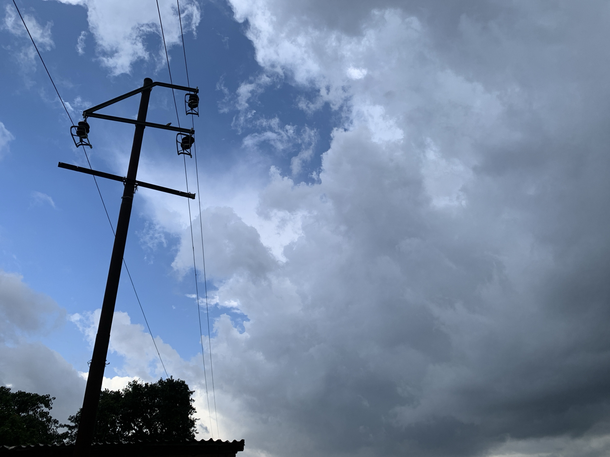 This image captures a utility pole silhouetted against a dramatic sky. The pole, a dark vertical element on the left side of the frame, supports multiple wires and insulators. It appears to be a power or telephone pole. In the foreground, at the very bottom, a dark, corrugated roofline and the dark silhouettes of trees are visible, suggesting a built-up area, possibly a residential or semi-rural setting.
The sky is the dominant feature, filled with a mix of blue sky and heavy, dark clouds, indicating dynamic weather conditions, perhaps approaching rain or a storm. Patches of brighter blue peek through in the upper left, contrasting with the ominous, gray masses that cover a large portion of the frame. The lighting suggests it is likely daytime, but the presence of dark clouds creates a moody atmosphere. There are no visible people or text in the image. The scene evokes a sense of anticipation, perhaps due to the impending weather. The location context of Kuru Karama, Nigeria, places the image in a specific geographic setting, but no direct visual cues within the image itself confirm this.