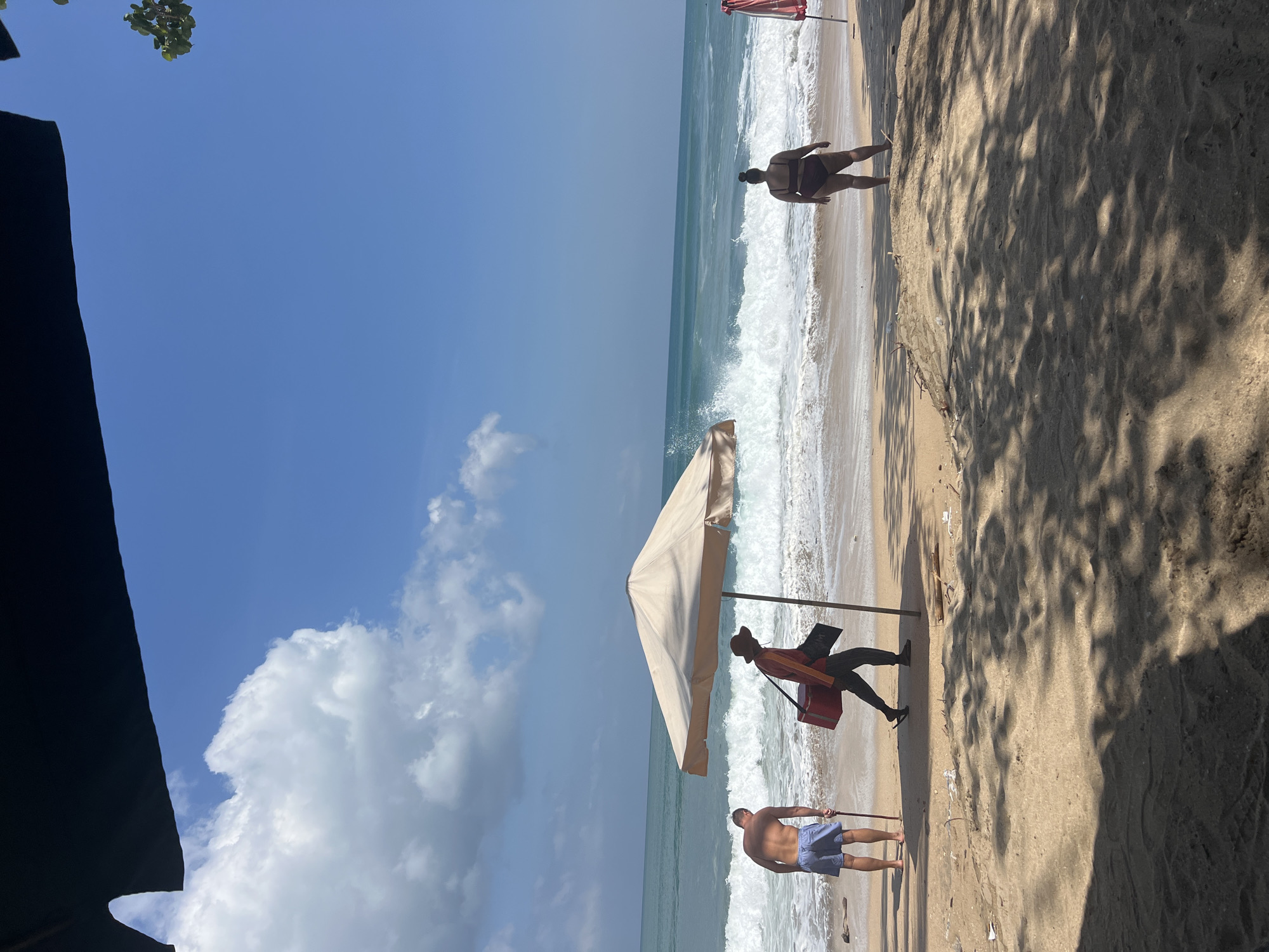 A bright, sunny day at Kuta Beach, Indonesia, is depicted in this image. The turquoise ocean churns with white-capped waves, meeting a sandy shore scattered with debris and vegetation. The foreground is dominated by the dappled shade of unseen trees, casting intricate patterns on the sand.
In the middle ground, three individuals are present. On the left, a man with a bare torso and blue shorts stands facing the ocean, holding a long stick. To his right, another person walks away from the viewer, wearing a hat and carrying a red cooler bag and a black flat object. Further to the right, a person in a dark bikini faces the sea, their back to the camera. An open beach umbrella provides shade for a portion of the sand. In the background, a red and white umbrella is visible. The sky is a vibrant blue, dotted with white, fluffy clouds.