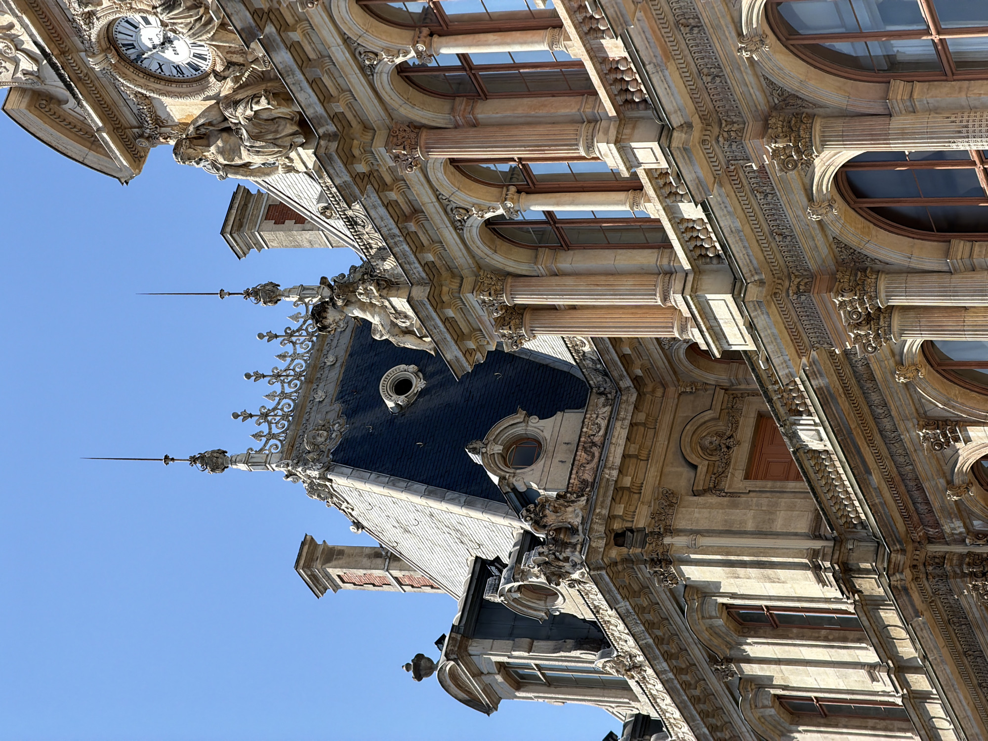 This image shows a detailed view of the Hôtel de Ville (City Hall) in Lyon, France, under a clear blue sky. The architecture is classical, characterized by ornate stonework, decorative sculptures, and a large clock face on the upper facade. The building features multiple levels with arched windows, Corinthian columns, and elaborate friezes. A steeply pitched, dark roof with dormer windows and decorative spires is visible. The scene captures the grandeur and historical significance of the building's exterior.