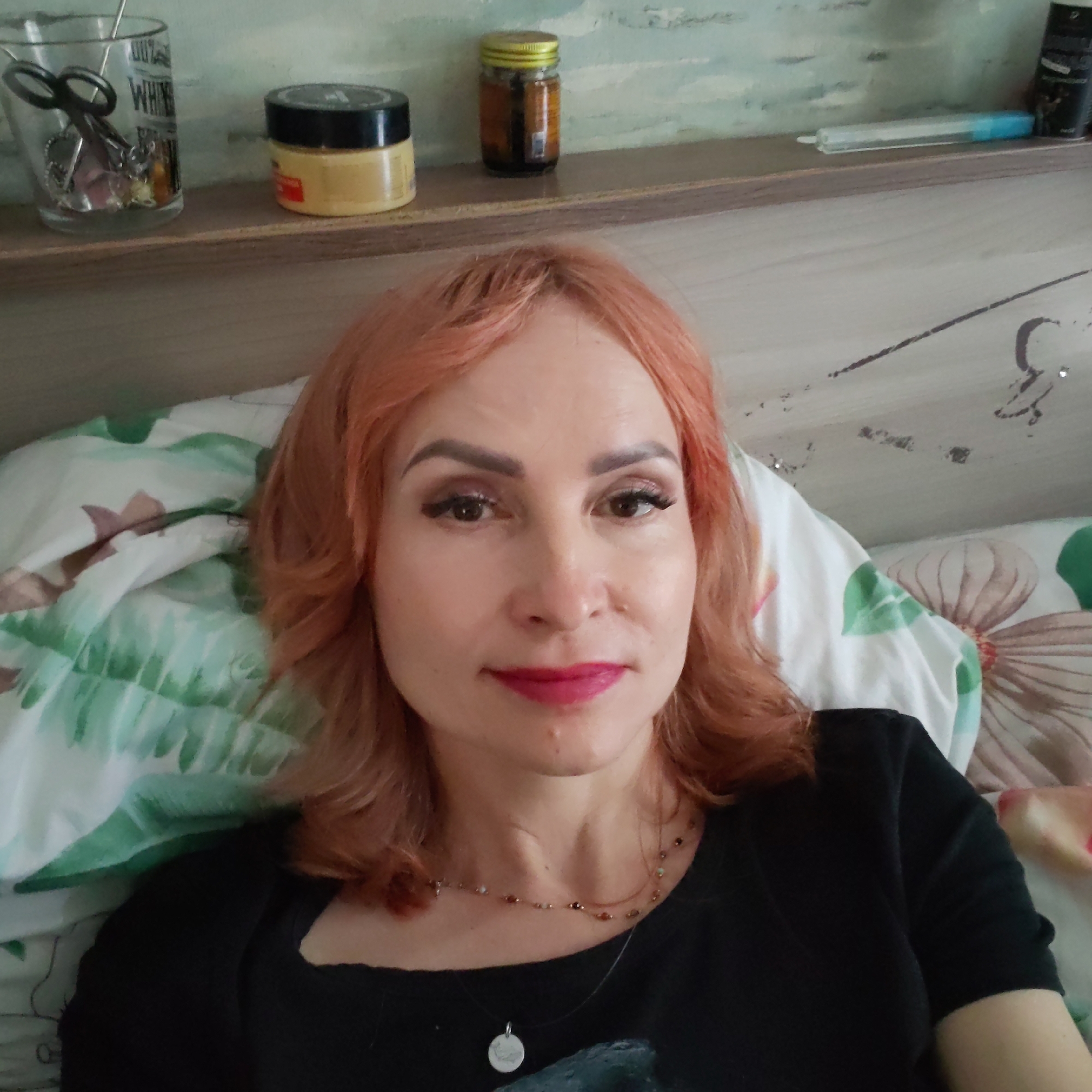 A woman with reddish-orange hair is lying in bed. She is wearing a black shirt, a necklace, and makeup. Behind her is a headboard with a shelf on which are several items, including a glass filled with scissors and other small metal objects, a yellow jar with a black lid, and a small glass jar with a yellow lid. The pillow and blanket are covered with a floral pattern. The image was taken indoors. There is no indication of a location in Chaikovsky, Russia.