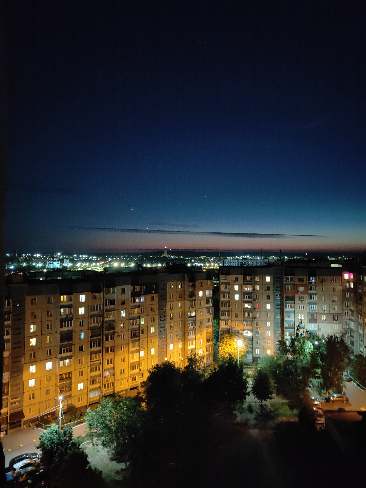 The image captures a nighttime view of a city from a high vantage point, likely an apartment building in Kryvyi Rih, Ukraine. The sky is a deep, dark blue, transitioning to a lighter hue near the horizon where a faint band of light suggests either the very early morning or late evening twilight. Several stars are visible in the upper portion of the sky, and two brighter celestial bodies, possibly planets, are also present.
Below the sky, a distant cityscape is illuminated by numerous lights. These lights appear as clusters and scattered points, indicating a residential and possibly industrial or commercial area. A thin, horizontal line of lighter color in the distance may be a cloud formation or atmospheric haze.
The foreground is dominated by the facade of a large, multi-story apartment building. The building's windows are lit from within, showing a pattern of yellow and occasional red lights, suggesting occupied apartments. The building's structure is visible, with balconies and the grid-like arrangement of windows.
In front of the apartment building, trees are silhouetted against the faint light. Some of these trees are densely leaved and dark, while others appear more sparse. A streetlamp is visible, casting a yellow glow on its surroundings and illuminating a patch of the ground and nearby vegetation.
At the base of the building and slightly to the left, several cars are parked. They are dark shapes in the low light, with some car lights visible. A road or pathway is also discernible, with a few more vehicles further into the scene. The ground area appears to be a courtyard or parking lot. The overall impression is one of urban living at night, with the glowing windows of apartments and the distant city lights creating a sense of a sleeping but active city.