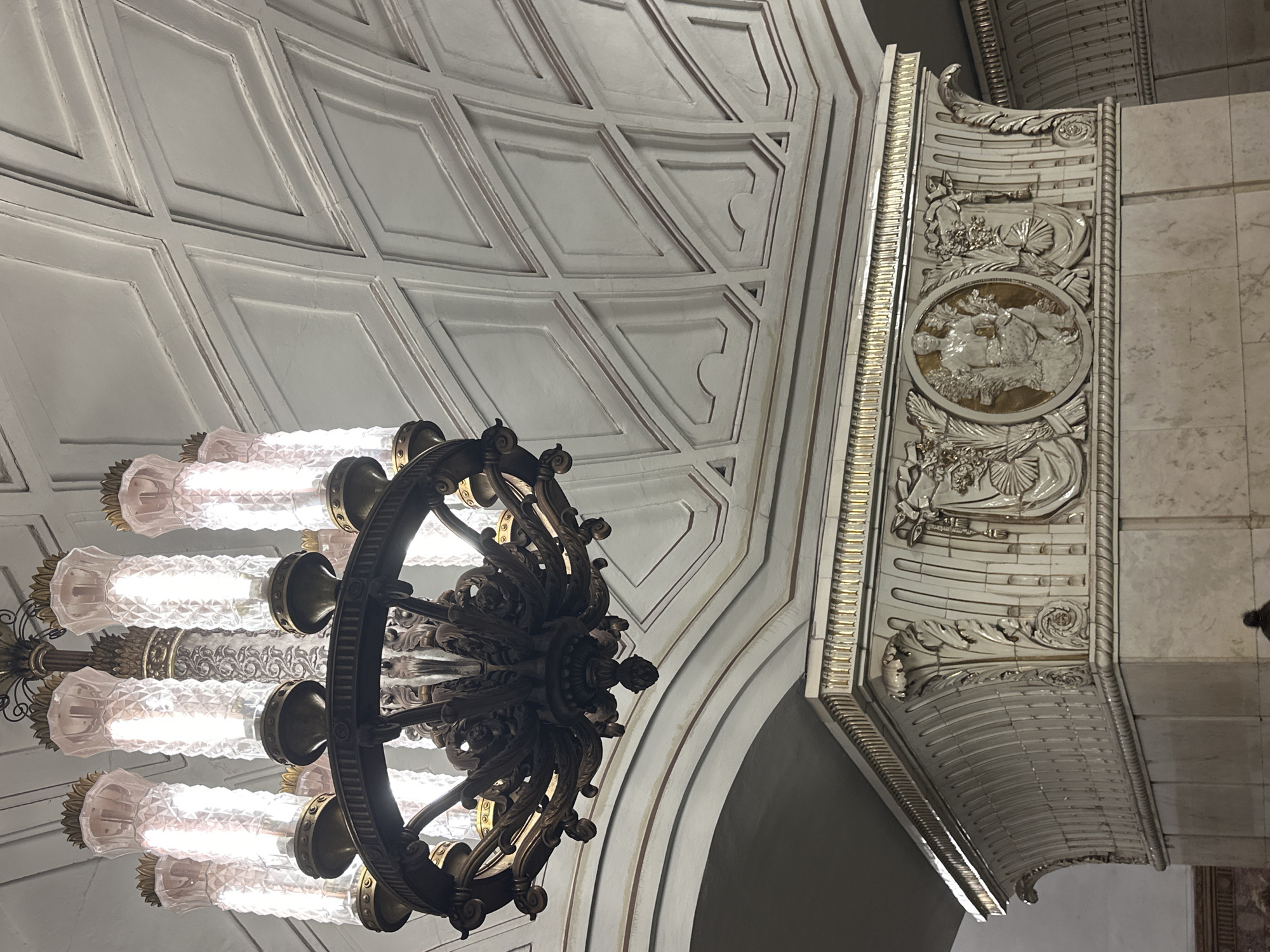 The image shows a grand chandelier with multiple light fixtures suspended from a decorative, vaulted ceiling. The ceiling features a patterned design of recessed panels outlined in thin, dark lines. Below the ceiling and to the right, a section of the ornate column is visible. This column is clad in white tiles with intricate relief carvings, including a central oval medallion with a sculpted figure. The column also displays decorative gold trim along its top edge. The overall aesthetic suggests an interior of a public building, likely a metro station or grand hall, given the architectural style and ornate decorations. This specific type of architectural style and the presence of such chandeliers are characteristic of Moscow's ornate metro stations, particularly those dating from the Soviet era.