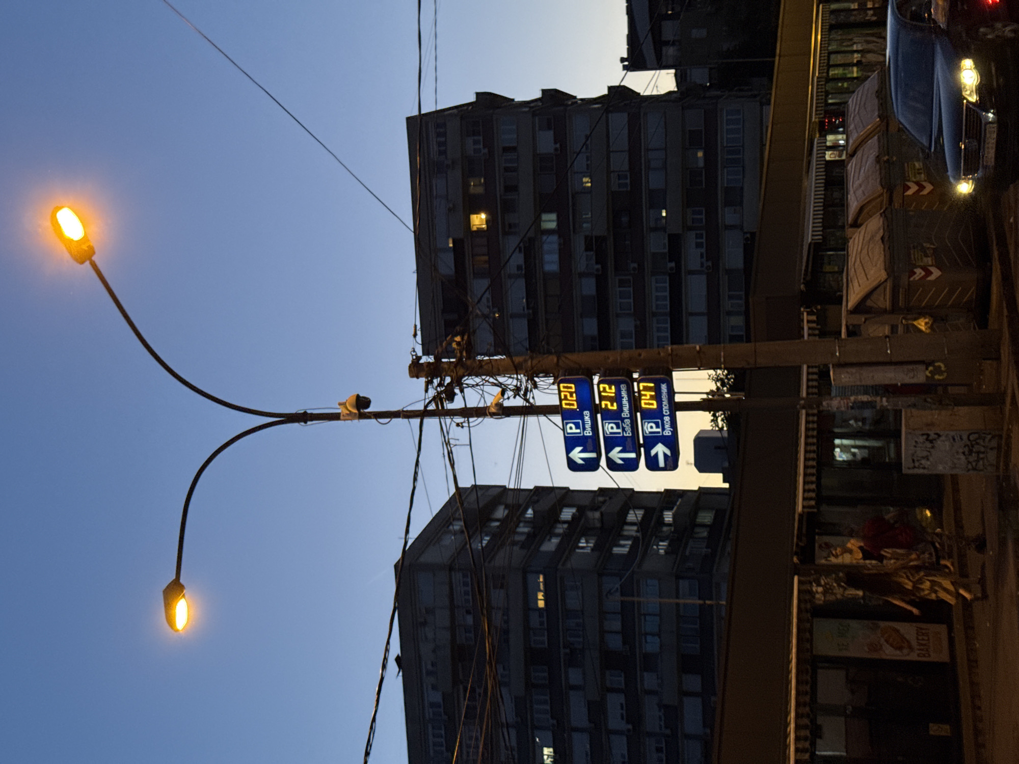 This image captures an urban street scene at dusk in the Vracar Urban Municipality, Belgrade, Serbia. The sky transitions from a deep blue overhead to a lighter, warm orange-yellow near the horizon line. Artificial lighting is prominent, including streetlights, illuminated windows in buildings, and vehicle headlights.
A central, tall utility pole dominates the midground, laden with numerous tangled black wires and equipped with at least two surveillance cameras, one yellow and one dark grey. Attached to this pole are three vertical digital parking signs displaying real-time parking availability and directional information in Cyrillic. The top sign, with an upward arrow, shows "P 020 Вишка" (P 020 Viška). The middle sign, also with an upward arrow, indicates "P 212 Баба Вишњина" (P 212 Baba Višnjina). The bottom sign, with a rightward arrow, displays "P 047 Вуков споменик" (P 047 Vukov spomenik). The names "Baba Višnjina" and "Vukov spomenik" confirm the location within Vracar.
Two large, multi-story buildings, likely residential or mixed-use, form the background. The building on the left is prominent, featuring numerous windows, some with warm interior lighting and visible air conditioning units. Below these buildings, at street level, are single-story storefronts with awnings, one of which has a visible "BAKERY" sign.
On the sidewalk in the lower left, two individuals are walking. One person with long hair, wearing a light-colored garment, is moving from left to right.
In the lower right foreground, a dark-colored car with illuminated headlights faces the viewer. To its right, a portion of a red vehicle is visible. Three large, rectangular, dark-colored industrial waste bins with orange and white markings are situated on the sidewalk behind the car. One bin has a yellow plastic bag hanging from it. The base of the utility pole exhibits graffiti. A dual-arm streetlight with two glowing yellow-orange lamps curves from the central pole in the upper left, providing street illumination.