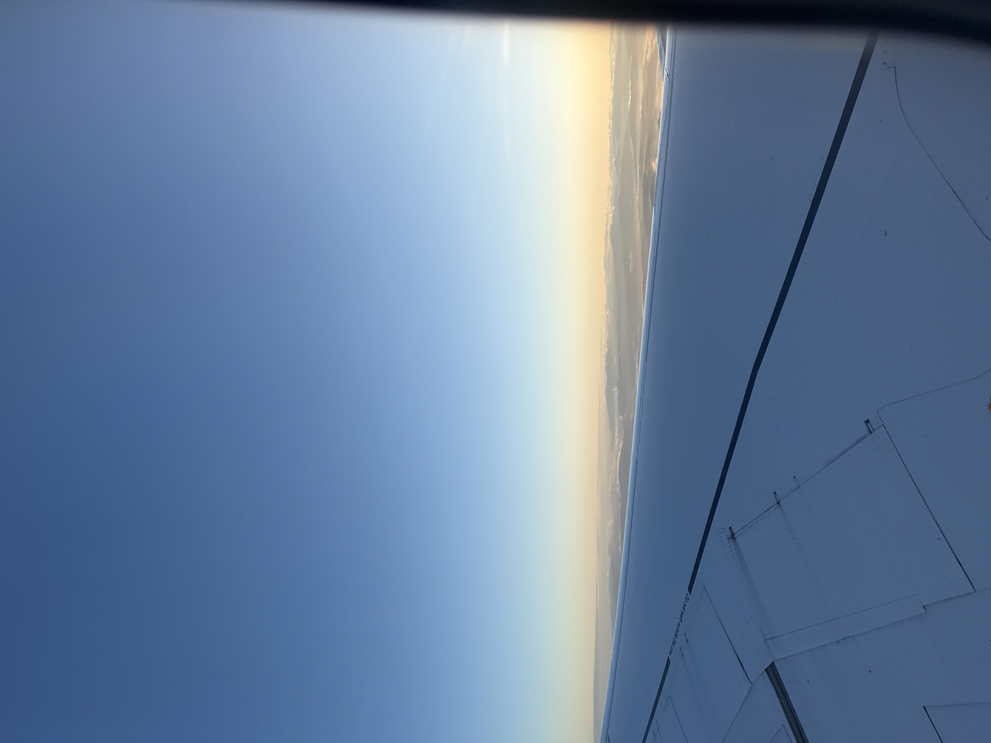 The photograph was taken from inside an airplane, looking out of a window and down at the ground. The lower right of the frame is filled by a portion of the airplane's wing, which appears to be white or light gray in color. Visible are panel lines and small rivets on the wing's surface. The sky is visible on the left side of the frame, transitioning from blue to a lighter yellow near the horizon. Along the horizon, there is a layer of haze or thin clouds giving a soft appearance to the area where the sky meets the earth. The location is possibly above Moreira, Portugal, which cannot be fully determined by the image provided.