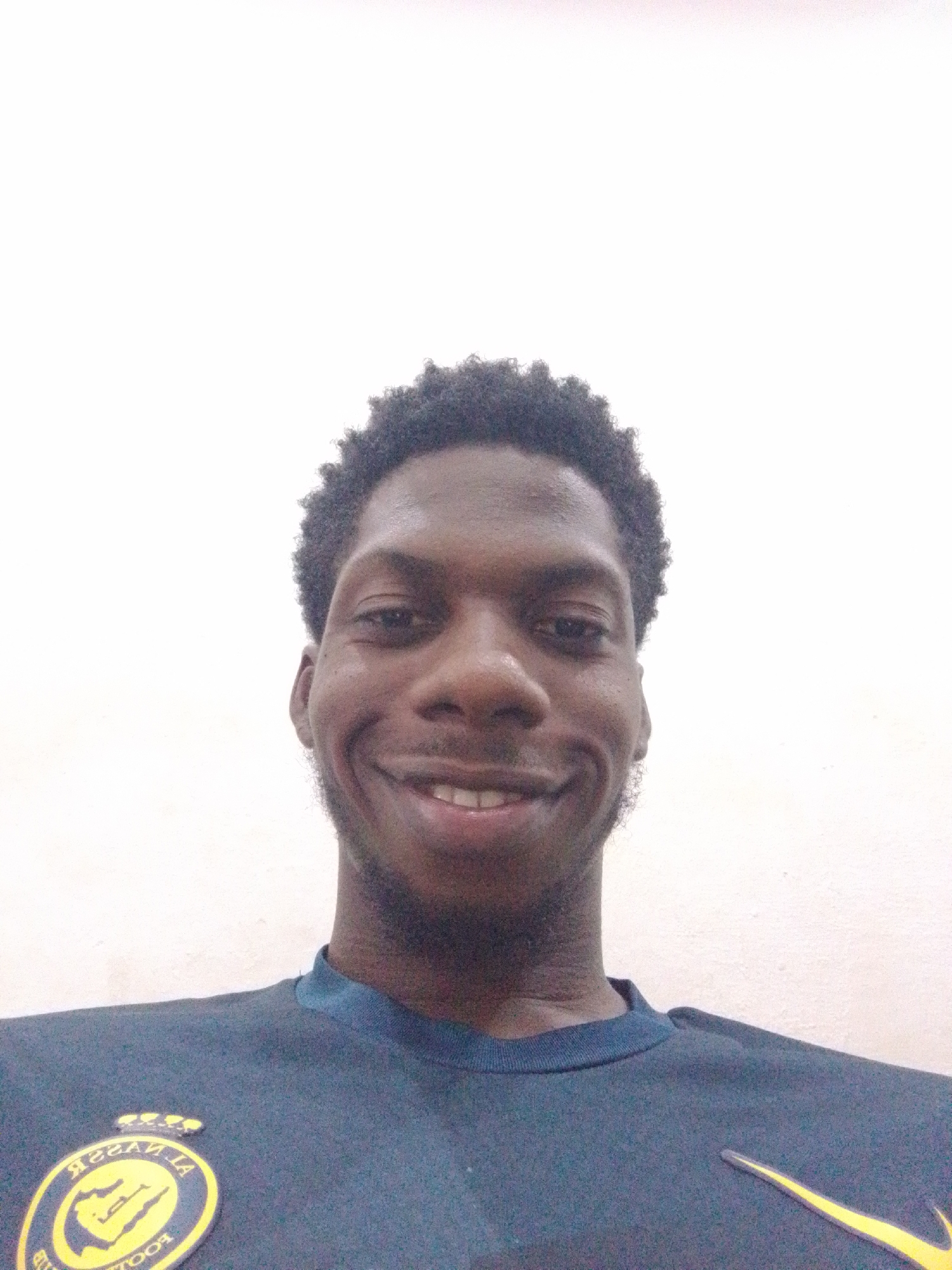 A close-up, selfie-style shot features a young Black man with dark, curly hair smiling broadly against a plain white background. He is wearing a dark blue t-shirt with a yellow and gold emblem on the left chest and a yellow Nike swoosh logo on the right side of the chest. The emblem appears to be circular with Arabic writing and a depiction of a camel and palm trees. The man's face is well-lit, with his teeth visible in his open-mouthed smile. His skin tone is dark brown, and he has a short beard. The image is cropped to focus on his upper body and face, with the background being mostly out of focus and brightly lit, suggesting an indoor setting. There are no discernible elements within the image that would indicate the specific location within Abeere, Nigeria.