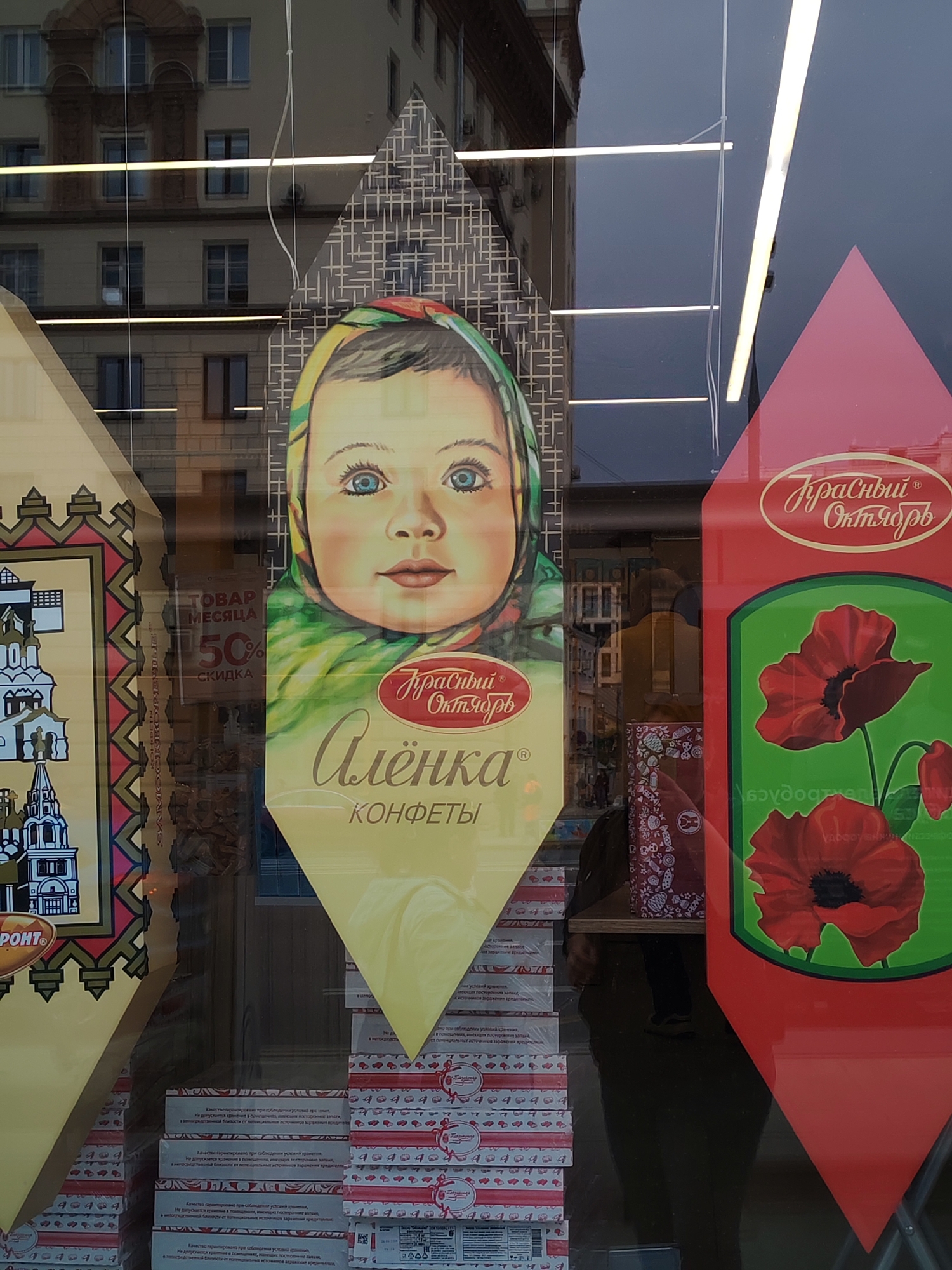 The image captures a storefront window in Moscow, Russia, featuring promotional displays for Russian confectionery brands, prominently "Krasny Oktyabr" (Red October). Three large, shaped cardboard cutouts are displayed on the window, partially obscuring the reflections behind them.
The central cutout is a vertical, diamond-shaped piece showcasing the iconic "Alenka" (Alyonka) candy design: a young baby with blue eyes and a colorful headscarf. Below the baby's portrait, the brand name "Красный Октябрь" (Red October) and "Алёнка КОНФЕТЫ" (Alyonka Candies) are visible in Cyrillic script.
To the left, another decorative cutout features a stylized illustration of a multi-domed church or cathedral, with text that includes "ФРОНТ" (Front), likely another candy series. On the right, a red, pointed cutout displays a vibrant green panel with red poppies, also branded with "Красный Октябрь" (Red October).
Reflected in the window are elements from both inside and outside the store. A multi-story building with numerous windows and architectural details, possibly an apartment complex or office building, is clearly reflected across the top and middle sections, suggesting a daytime setting, potentially overcast. Inside the store, horizontal fluorescent strip lights are reflected, along with stacks of candy boxes on shelves. A sign inside the store reads "ТОВАР МЕСЯЦА 50% СКИДКА" (PRODUCT OF THE MONTH 50% DISCOUNT), indicating a sale. Partial reflections of a person in dark clothing are also visible in the lower right, suggesting a passerby or someone inside. The small text on the candy boxes mentions "Качество гарантировано при соблюдении условий хранения" (Quality guaranteed if storage conditions are met).