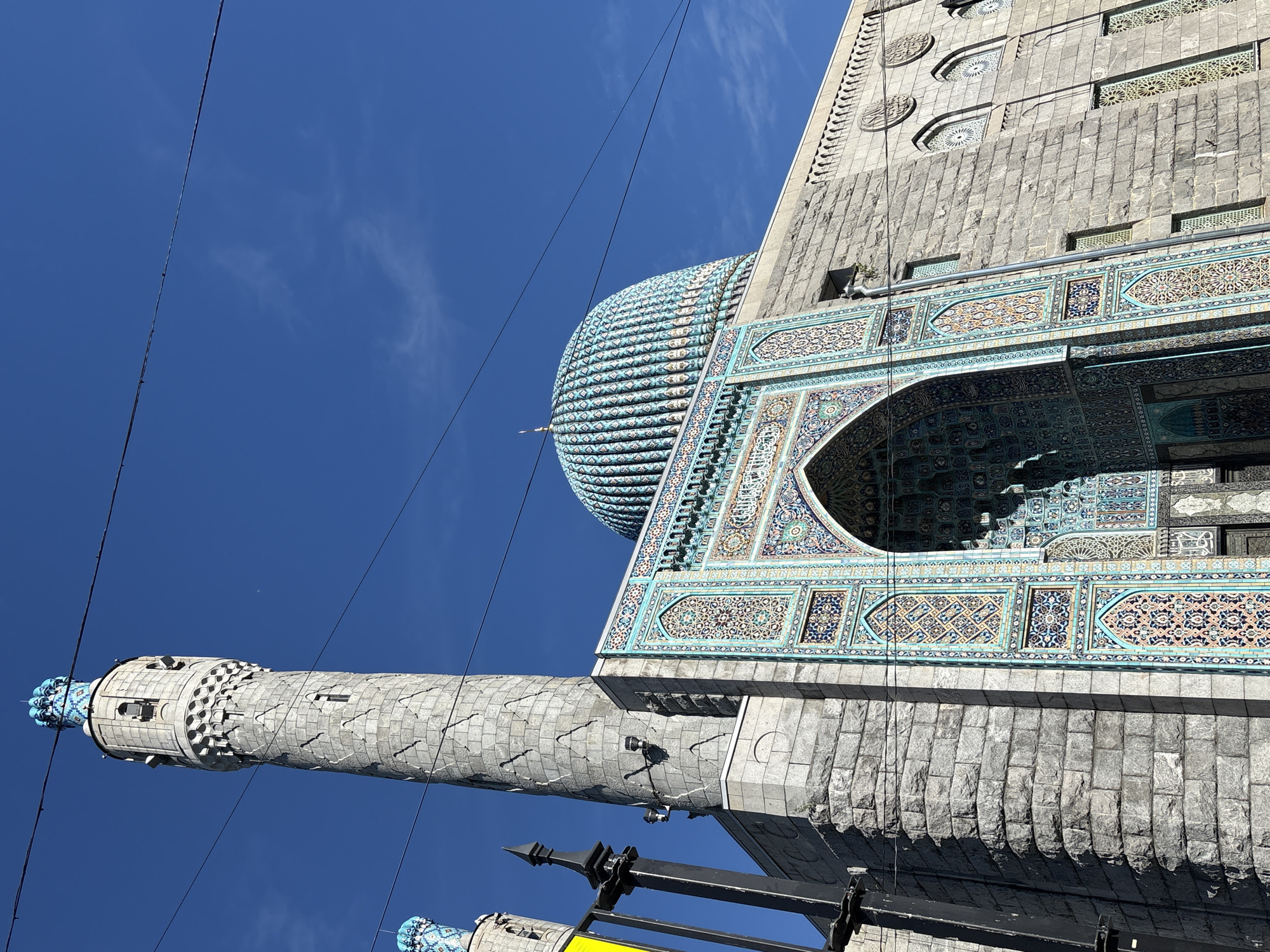 The image shows the Saint Petersburg Mosque, located in Saint Petersburg, Russia. The mosque features a striking blue tiled dome and intricate decorative tiling around its arched entrance. A tall, cylindrical minaret, constructed from stone, rises to the left of the main building. The architecture exhibits Islamic design elements, with patterns and calligraphy visible on the facade. The sky above is a clear, bright blue, with a few faint wisps of clouds. Power lines are visible crossing the upper portion of the frame. The building is constructed of rough-hewn stone blocks. The image is taken from a low angle, emphasizing the grandeur of the mosque.