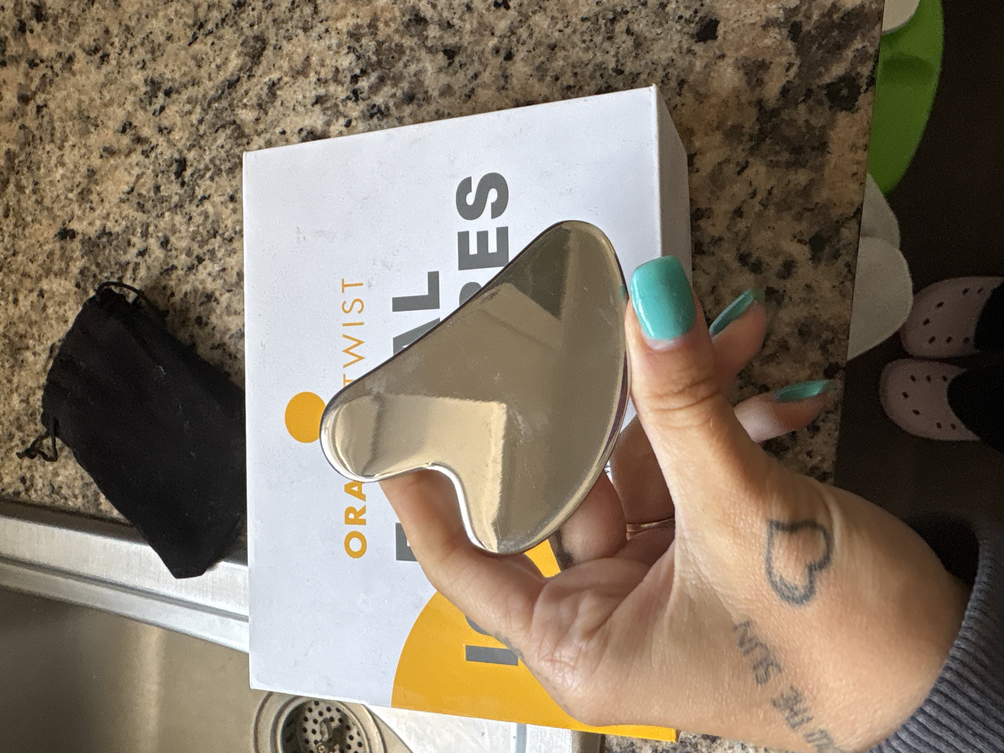 A close-up, high-angle photograph shows a human hand, adorned with vibrant teal nail polish, holding a reflective, silver-toned, heart-shaped gua sha tool. The back of the hand displays two tattoos: a black outline of a heart and partially visible script that reads "THE SUN" (reversed). A thin gold-colored ring is visible on one of the fingers.
The gua sha tool is held over a white product box featuring an orange circular logo and text. Visible portions of the text include "ORANGE TWIST" on the side and larger letters "AL ES" on the top surface. A black drawstring pouch lies to the left of the box.
The objects rest on a speckled, light brown granite-like countertop. A stainless steel kitchen sink with a drain is visible in the lower left quadrant of the frame. In the background, a pair of light pink clogs and a green object are partially visible. The scene is an indoor setting, likely a kitchen or utility area, under bright, diffused lighting. The photo was taken in Seal Beach, United States of America.