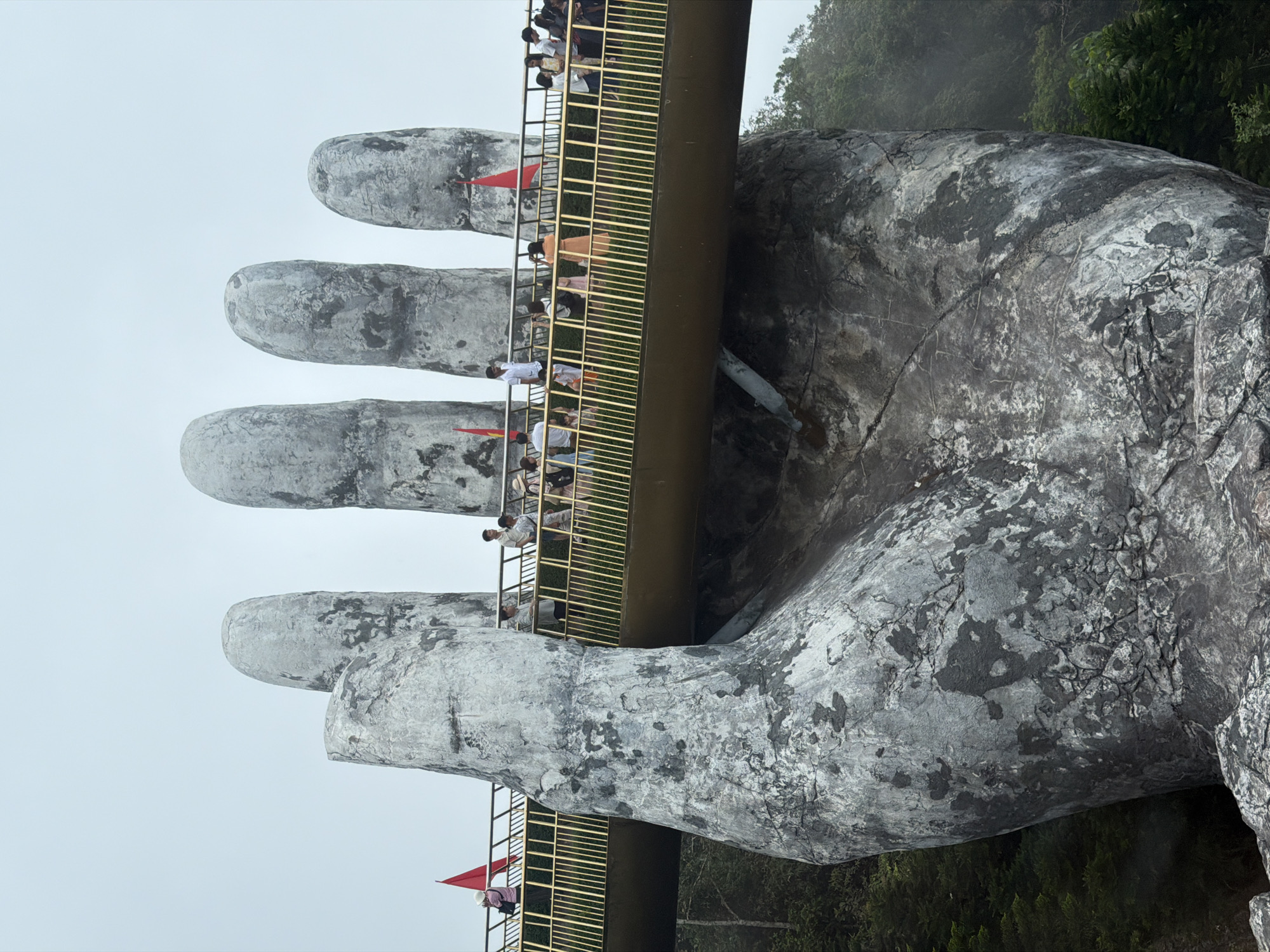 This image depicts the iconic Golden Bridge (Cầu Vàng) in Đà Nẵng, Vietnam, showcasing its unique architecture. The central subject is a massive, weathered hand sculpture, seemingly crafted from concrete or stone, emerging from the landscape to cradle a pedestrian bridge. The hand exhibits a rugged texture with variations in grey and dark tones, suggesting an aged or natural stone appearance.
The bridge features prominent golden-colored railings with a horizontal grid pattern, contrasting sharply with the colossal grey hand and the muted sky. Numerous individuals, likely tourists, are seen walking along the bridge, some standing and observing, others moving along its path. They are dressed in a variety of casual clothing, indicating a leisurely sightseeing activity.
The background is dominated by lush, dark green foliage, indicative of a mountainous or forested environment, which is partially obscured by a soft mist or haze. The sky appears overcast, creating a diffused light that lends an atmospheric, almost ethereal quality to the scene. Small red flags are intermittently attached to the golden railing of the bridge, adding minor splashes of color. The image captures the bridge as a bustling tourist destination amidst a serene, misty landscape. There is no visible text in the image.