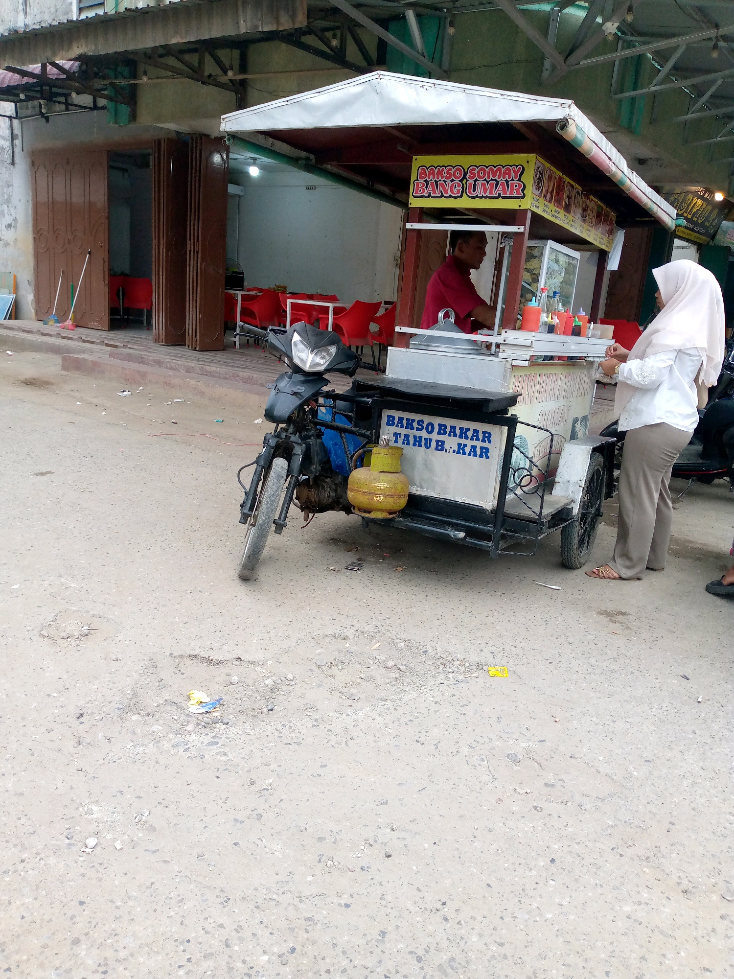 In Keudegeudong, Indonesia, a street scene unfolds with a food cart selling "Bakso Bakar Tahu Bakar" (grilled meatballs and grilled tofu). The cart, attached to a motorcycle, is positioned on a dirt and asphalt road. A gas cylinder is visible on the cart. Behind the cart, a food stall with a red-clad vendor is visible, and customers are seated at red chairs. To the left, a building with brown sliding doors and brooms suggests commercial activity. A woman wearing a white hijab and a white top stands near the food cart. The environment appears to be an outdoor urban setting with some debris on the ground.