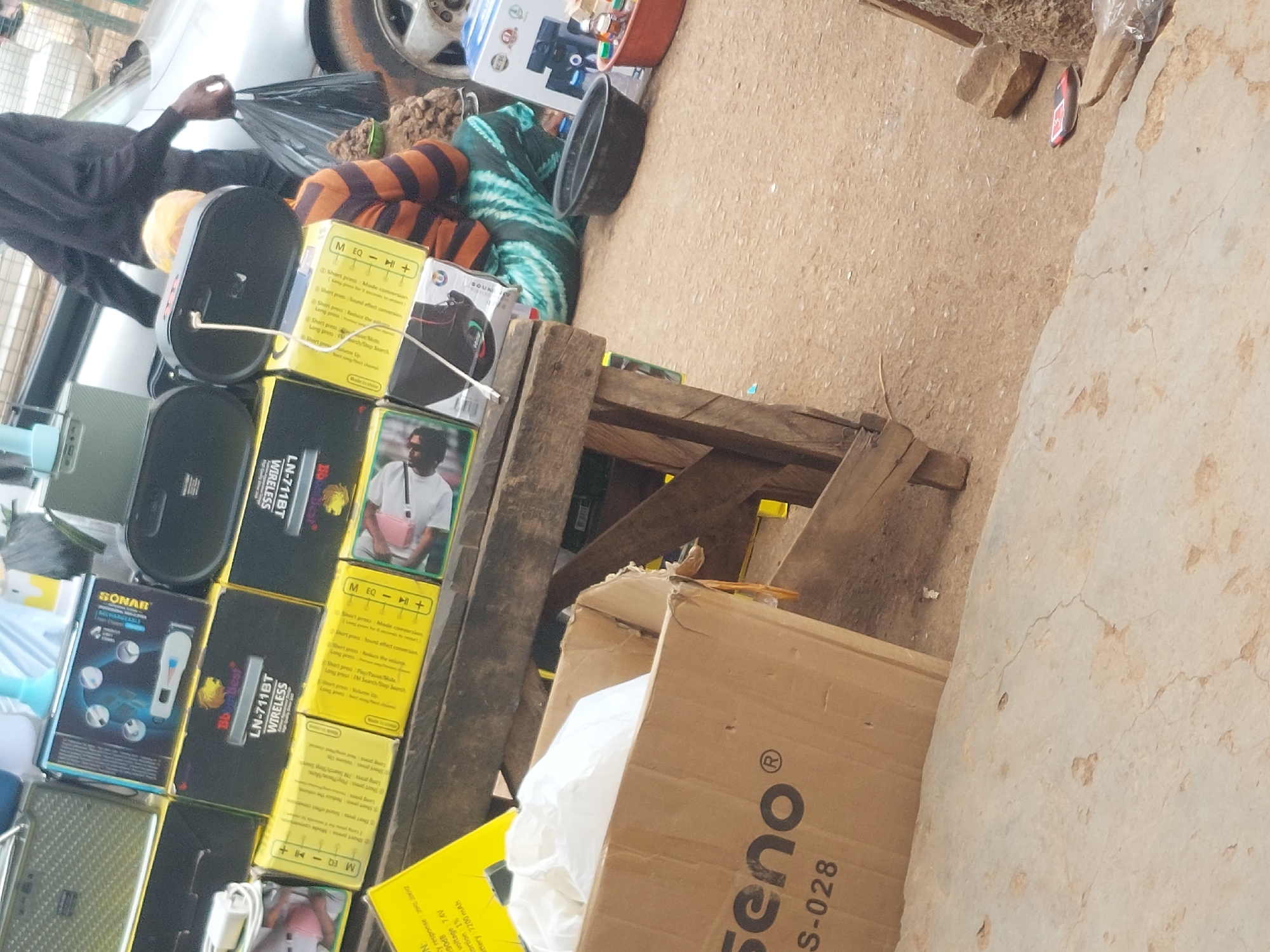The image captures an outdoor market or roadside stall at an unspecified location in the city of Unknown, Unknown. In the immediate foreground, a partially open cardboard box, labeled "Seno® S-028," rests on a light-colored, cracked concrete surface, containing crumpled white material.
The central feature is a rustic wooden stall displaying an assortment of packaged electronic devices. The stall is constructed from rough planks of wood. Stacked upon it are numerous yellow and black cardboard boxes, primarily for "LN-711BT WIRELESS" devices, prominently featuring a "Bluedest" lion logo. A smaller blue box on the upper stack is labeled "SONAR" and displays images of a compact digital recorder. Another yellow box, partially visible on the far left, lists specifications including "OUTPUT VOLTAGE 7.4V," "DISTORTION 1%," and "BATTERY 7200mAh." Several boxes depict individuals using headphones or holding mobile phones. On top of the stack, a black oval-shaped speaker is connected by a white cable, which trails downwards. A light green textured speaker and a light blue plastic object (possibly a small fan or speaker) are also on the stall.
To the right of the stall, a person is seated on the ground, their back partially obscured from view. They wear a long-sleeved shirt with horizontal orange and dark brown stripes and a lower garment or skirt in a turquoise and white patterned fabric. A light-colored head covering is visible.
Further in the background, a second person is standing, facing away from the viewer, clad in a dark (likely black) long-sleeved shirt and dark trousers. This individual holds a black plastic bag in their right hand and is positioned near the rear portion of a silver car, which is partially visible.
The ground surface transitions from the foreground concrete to a packed, light brown dirt or sandy area further back and to the right. On this dirt near the seated person, a black plastic bowl, a reddish-brown plastic bowl containing multiple small bottles, and several lumps of brownish, organic material (possibly roots or tubers) are arranged. A silver car wheel with a dirty tire is visible on the far right, and a red and black sandal or flip-flop lies on the dirt ground in the lower right background.