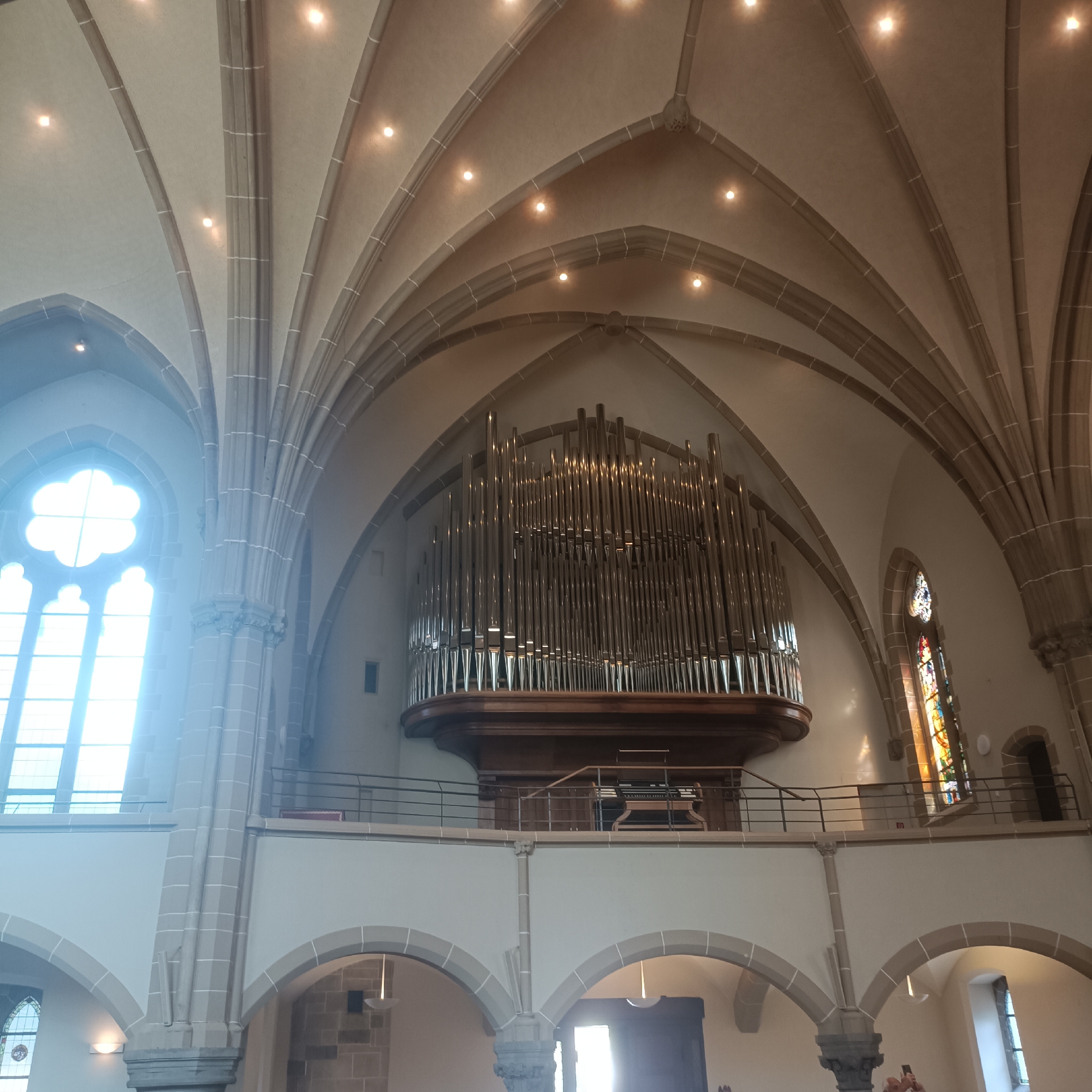 The image shows the interior of a church, likely in Bad Nauheim, Germany, featuring a grand pipe organ as the central focal point. The organ is mounted on a wooden structure above a balcony and consists of numerous metallic pipes of varying lengths and widths. The church's architecture is characterized by high, arched ceilings with stone ribs and a plastered surface, illuminated by recessed lights.
To the left, a large, decorative window with a cloverleaf-shaped upper section and arched lower panes is visible. Sunlight streams through this window, creating a bright, hazy effect. To the right of the organ, a stained-glass window with colorful patterns is partially visible.
The balcony below the organ is enclosed by a metal railing. Below the balcony, arched doorways or alcoves are present, with stone surrounds. The walls appear to be plastered and light-colored, contrasting with the darker stone elements. The floor is not visible.
No people are clearly identifiable in the image, although there is a suggestion of hands in the bottom right corner, possibly holding a phone. The overall atmosphere is one of solemnity and architectural grandeur.