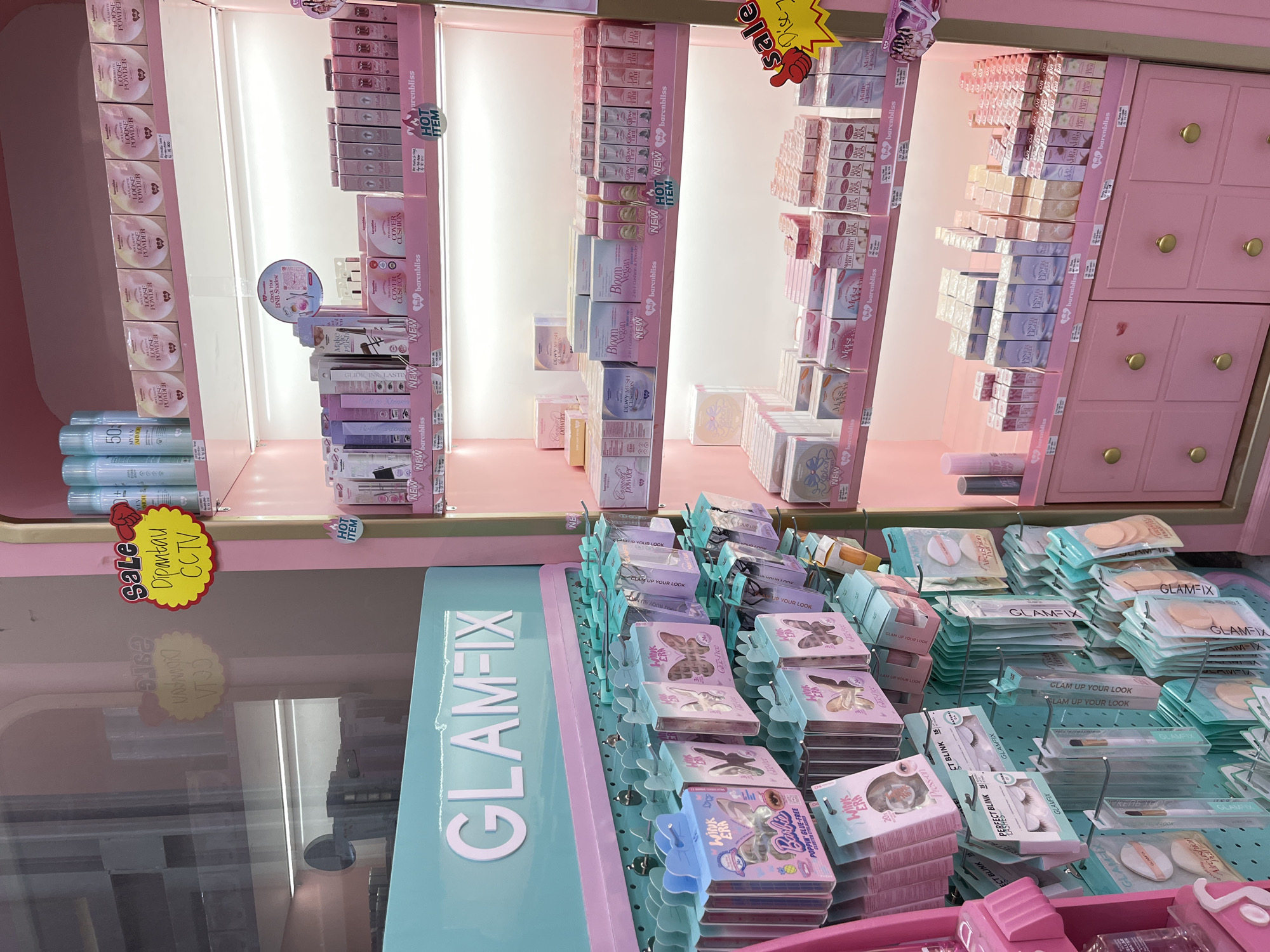 The image depicts the interior of a retail store, likely a beauty or cosmetics shop, predominantly featuring pink and teal color schemes. Located in Lhokseumawe, Indonesia, the store displays various beauty products arranged on shelves and pegboards. Prominent signage includes "GLAM-FIX" in large white letters on a teal background. Products are neatly packaged in boxes and blister packs, with brands such as "barenbliss" visible. Discounts or sales are advertised with a yellow sign reading "SALE" and "Dipantau CCTV," and another smaller red sign with "sale". The shelves are illuminated by bright lighting, highlighting the merchandise. The overall scene is clean and organized, typical of a retail environment. There are no people visible in the image.