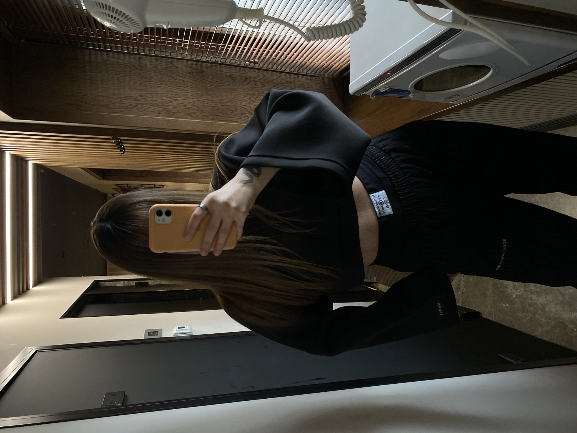 A person with long, brown hair is taking a photo of themselves in a mirror using a peach-colored phone. The person is wearing a black crop top and black pants. A hairdryer and a small white appliance are visible above the person's head. A dark doorway is visible to the left. The location is possibly a bathroom. The scene is unlikely to be in Votkinsk, Russia, given the details visible are not characteristic of that location.