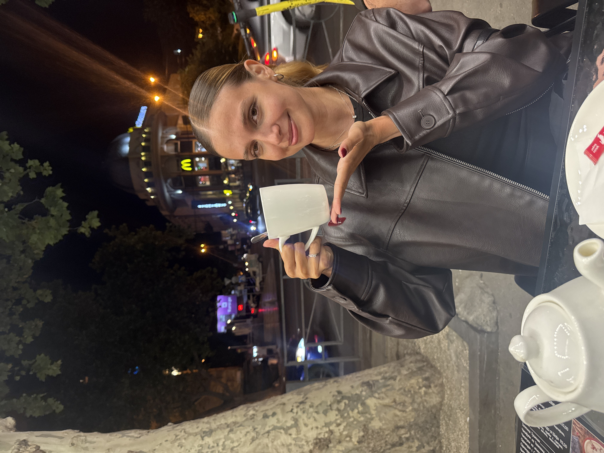 A young woman with blonde hair pulled back in a ponytail is sitting at an outdoor cafe at night. She is wearing a brown leather jacket and a black top. She is holding a white mug in her left hand and pointing at it with her right index finger. She is smiling at the camera. In the background, there are lights from buildings and streetlights, suggesting an urban setting. A white teapot and a menu are visible on the table in front of her. The menu has text in what appears to be a Cyrillic alphabet, and some English words like "White Sugar" are also visible. The overall scene suggests a casual evening outing.