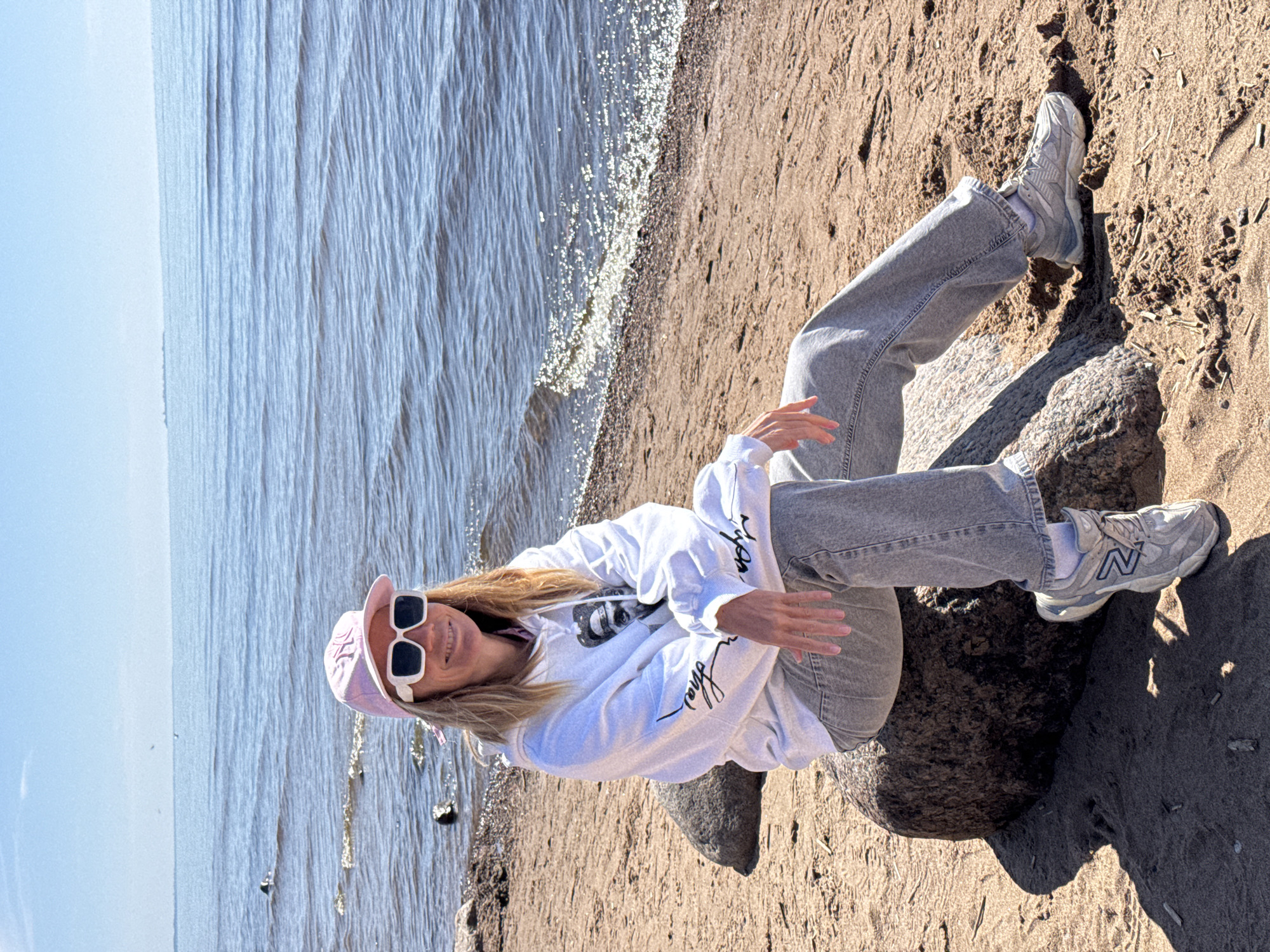 A young woman is sitting on a rock on a sandy beach by the sea in Komarovo, Russia. She is wearing a white hoodie, grey jeans, grey New Balance sneakers, a pink baseball cap, and white-rimmed sunglasses. The sea is to the left, with small waves lapping the shore. The sand stretches out around her, leading to a darker, rockier area to the right. The sky is light blue.