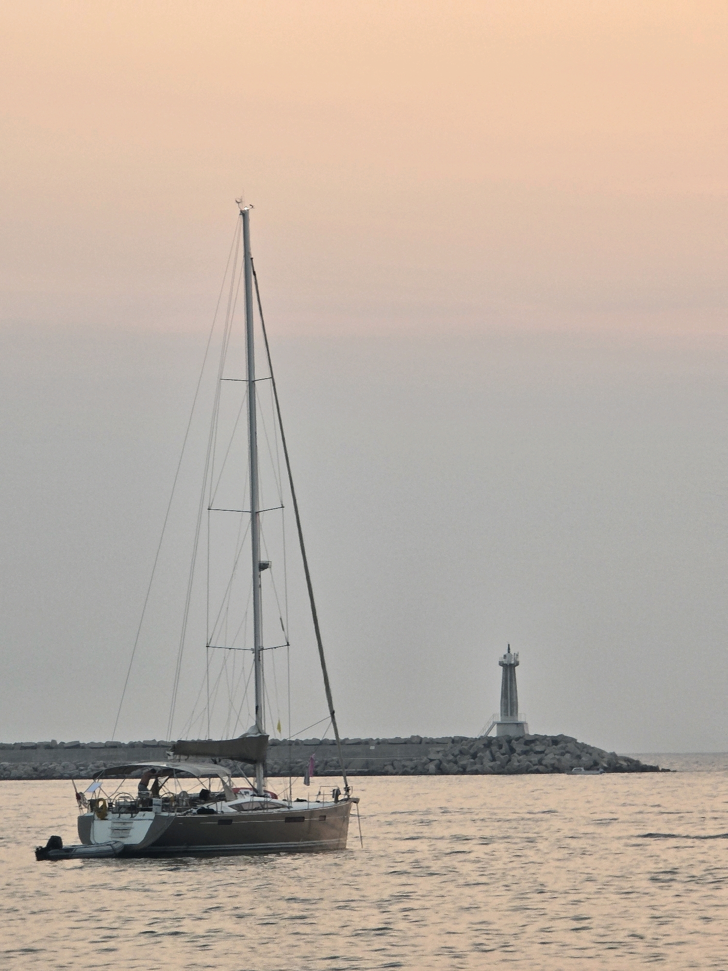 The image depicts a sailboat on the water, with a lighthouse situated on a rocky breakwater in the background. The sky is a soft, pastel hue, indicating either sunrise or sunset. The water is calm and reflects the colors of the sky.
The sailboat is positioned in the foreground, appearing to be either docked or slowly cruising. It is a modern vessel with a tall mast and rigging. Several people are visible on the deck of the sailboat, engaged in activities consistent with being on a boat.
In the mid-ground, a breakwater made of large stones extends into the water. Atop this breakwater stands a lighthouse, a tall, slender structure designed to guide ships. The lighthouse has a distinct lantern room at its peak.
The overall scene is serene and evokes a sense of maritime tranquility. Given the visual elements, this scene is likely located at the entrance to a harbor or bay, possibly near a coastal city. Without further context, it is impossible to definitively place this image in Bar, Montenegro, but such a landscape with a breakwater and lighthouse is plausible for a coastal city.