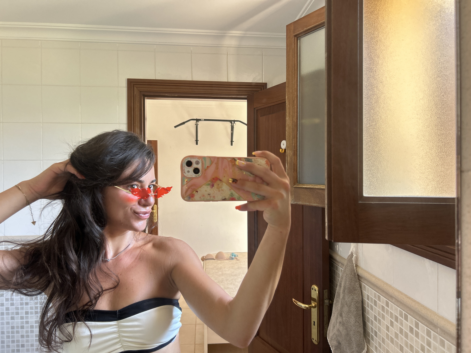 In the city of Capdepera, Spain, a woman takes a mirror selfie in what appears to be a bathroom. She is looking to her left, with her right hand touching her hair and her left hand holding a phone. She is wearing red, flame-shaped sunglasses and a white and black strapless top. The background shows tiled walls, a wooden door, and a pull-up bar mounted on the wall.