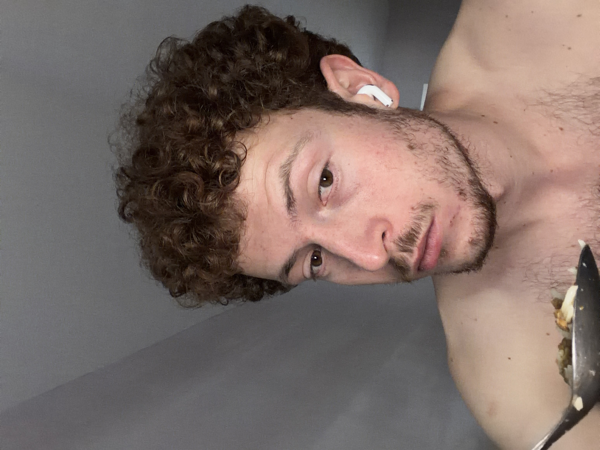 The image depicts a person with curly brown hair and a short beard, wearing an earbud in their left ear. They are holding a spoonful of food towards the camera. The person appears to be indoors against a grey wall. There is no information to discern the city or country.
