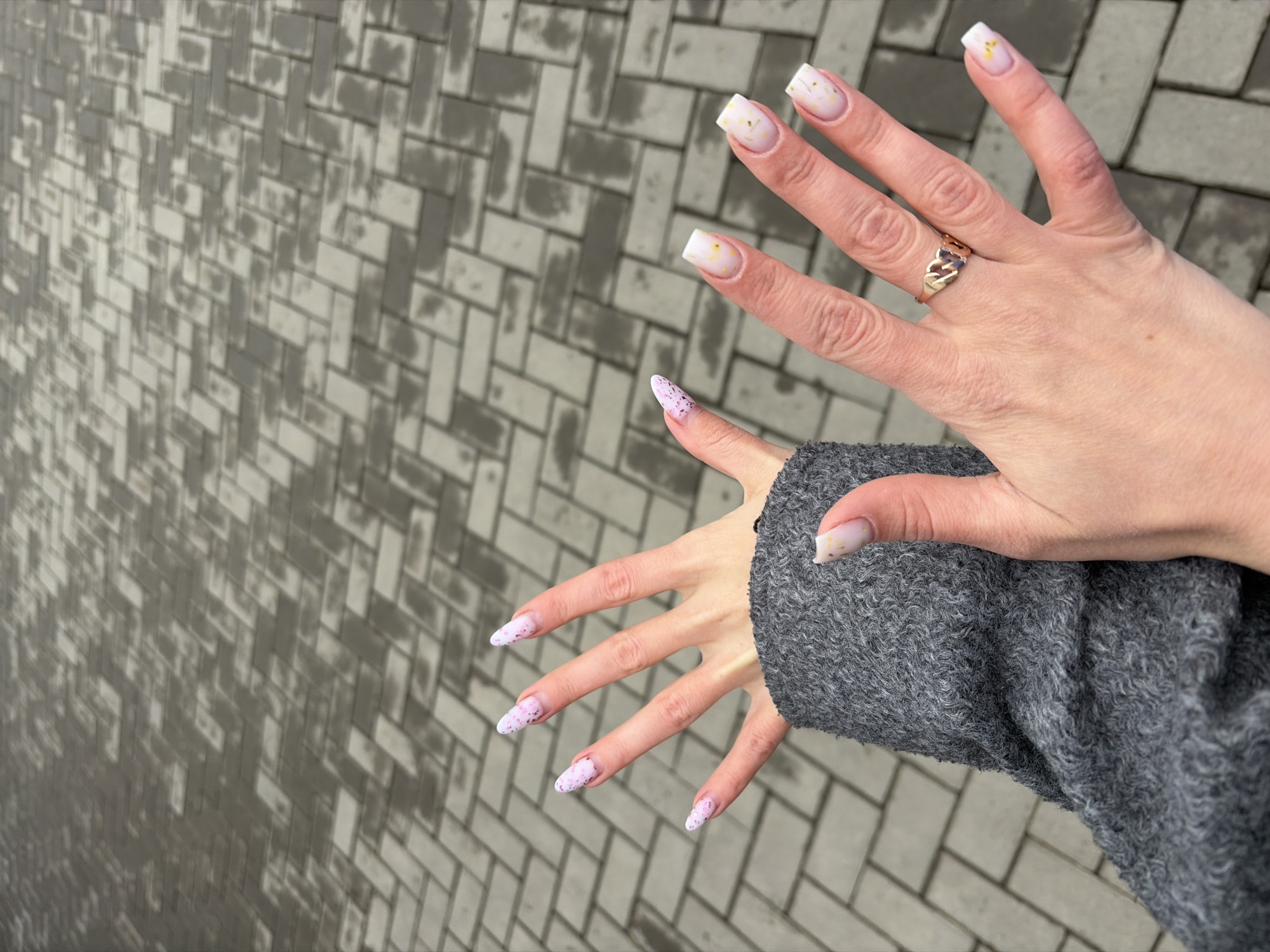 Two hands are displayed against a backdrop of a grey brick walkway. The upper hand, adorned with a gold ring on the ring finger, exhibits square-shaped nails with a white tip and gold flecks. The lower hand, partially covered by a grey sweater sleeve, shows almond-shaped nails with a white base and purple speckles. The location appears to be an outdoor urban area with distinct brickwork. The location is NOT identifiable.