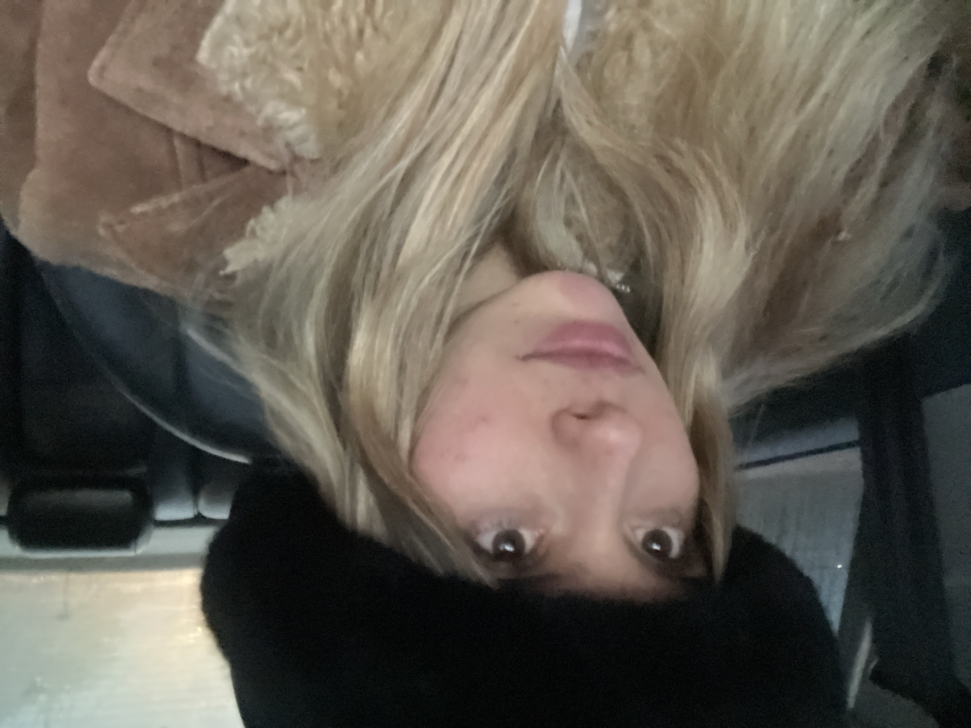 A woman with blonde hair is looking down at the camera. She is wearing a brown jacket with fur trim and a black fur hat. She appears to be indoors, possibly in a vehicle. The location is unknown, and there is no information to suggest this image was taken in San Fernando del Valle de Catamarca, Argentina.