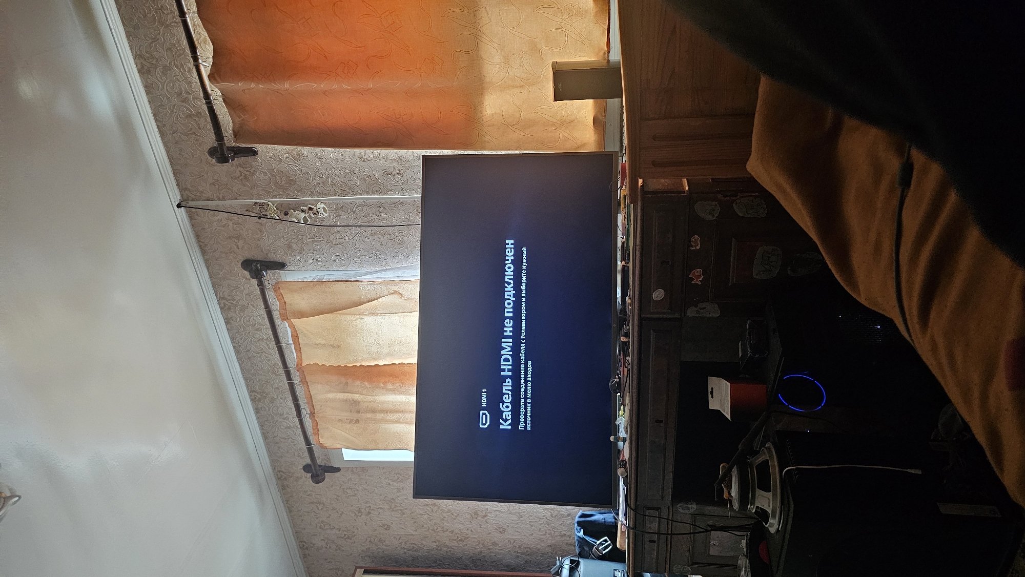 The image shows a television screen displaying a message in Russian: "HDMI cable is not connected. Check the cable connection to the TV and select the correct input source." The TV is placed on a wooden desk in a room with patterned wallpaper and orange curtains. There are various objects on the desk, including a speaker and what appears to be a computer tower. The overall setting suggests a domestic environment, possibly in the city of Nezhylovka, Russia. There are no people visible in the image.