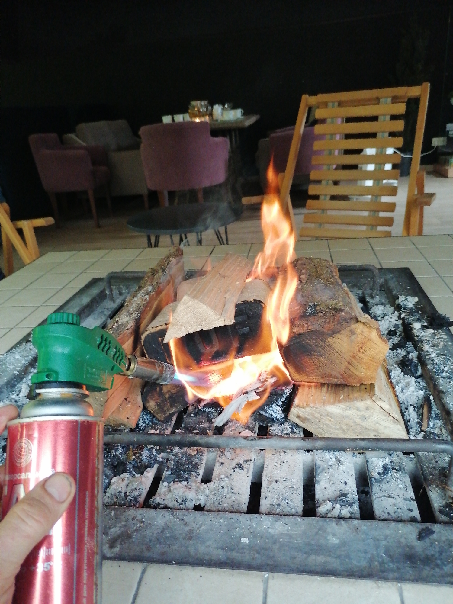 A close-up shot captures the moment a fire is being ignited or stoked in a fire pit. In the foreground, a hand holds a red gas canister with a green torch attachment, directing a bright blue and orange flame towards several wooden logs. The logs are resting on a metal grate, with significant ash accumulation beneath, indicating prior use of the fire pit. Bright orange and yellow flames rise from the burning wood, casting a warm glow.
The fire pit is set into a light-colored tiled surface. In the background, a lounge area is visible, dimly lit, suggesting evening or nighttime. This area features several upholstered armchairs, some in a deep purple hue and others in lighter grey or beige tones, arranged around small tables. A wooden slatted chair is prominently positioned on the right side of the frame, near the fire. The overall setting appears to be an outdoor patio or a covered, semi-outdoor area designed for relaxation.
Visible text on the red gas canister includes "Универсал" written in Cyrillic script, along with other smaller, partially obscured Cyrillic words and a globe-like logo. The scene conveys a cozy atmosphere, likely for warmth or ambiance.