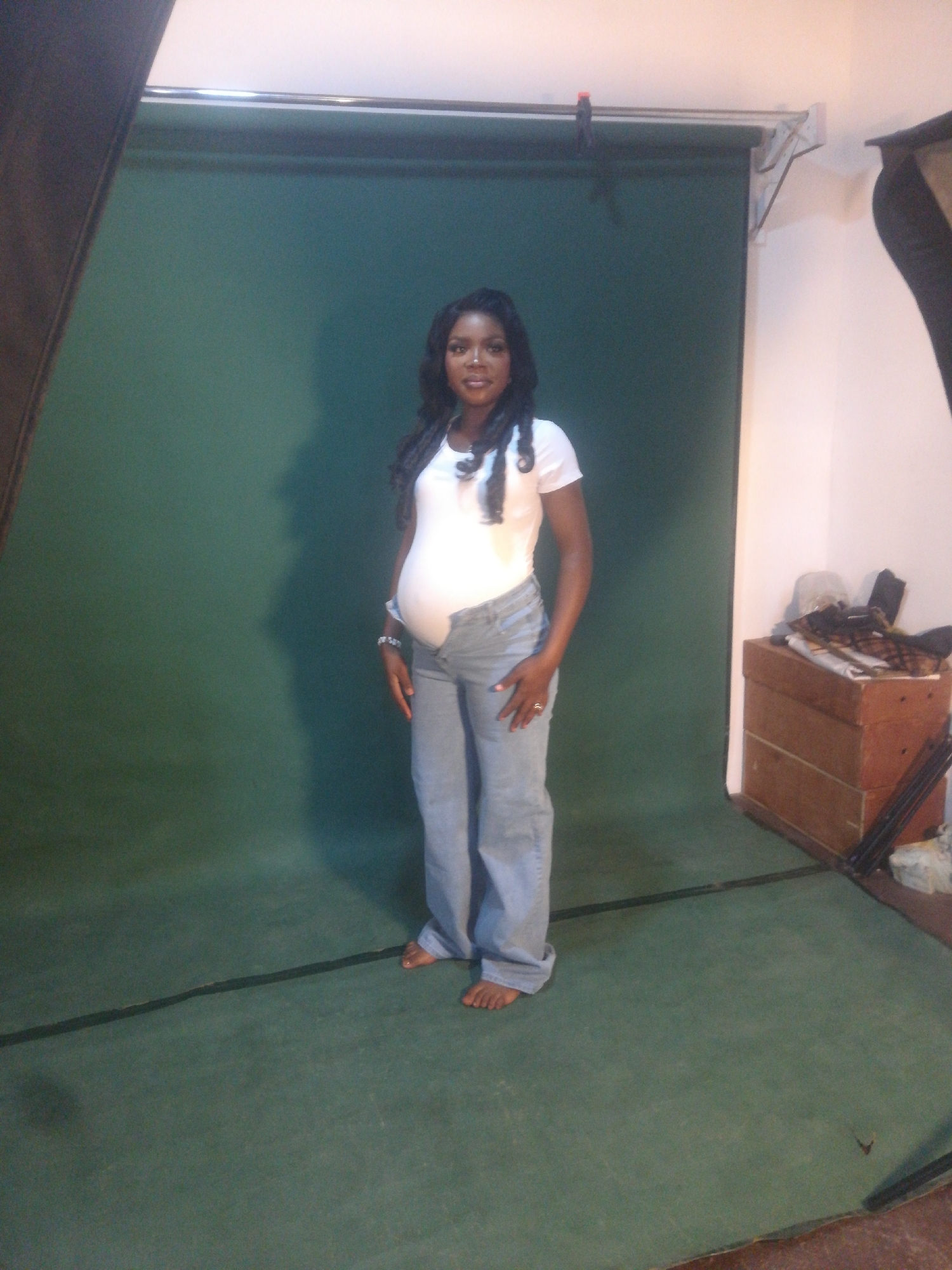 A full-shot, eye-level, indoor photograph captures a pregnant Black woman standing on a green floor in front of a dark green backdrop. The woman, who appears to be in her late 20s to early 30s, is facing the camera and smiling. She has long, dark, curly hair, and is wearing a white t-shirt and light blue jeans. Her left hand is resting on her hip, and she is wearing a ring on her left ring finger and a bracelet on her left wrist. The background is a plain, dark green fabric. To the right of the woman is a wooden chest or box, topped with various items, possibly clothing or fabric. The lighting in the scene appears to be artificial, casting a shadow behind the woman on the backdrop. The overall environment suggests a photo studio setting. The photograph was taken in an unknown location in an unknown city.