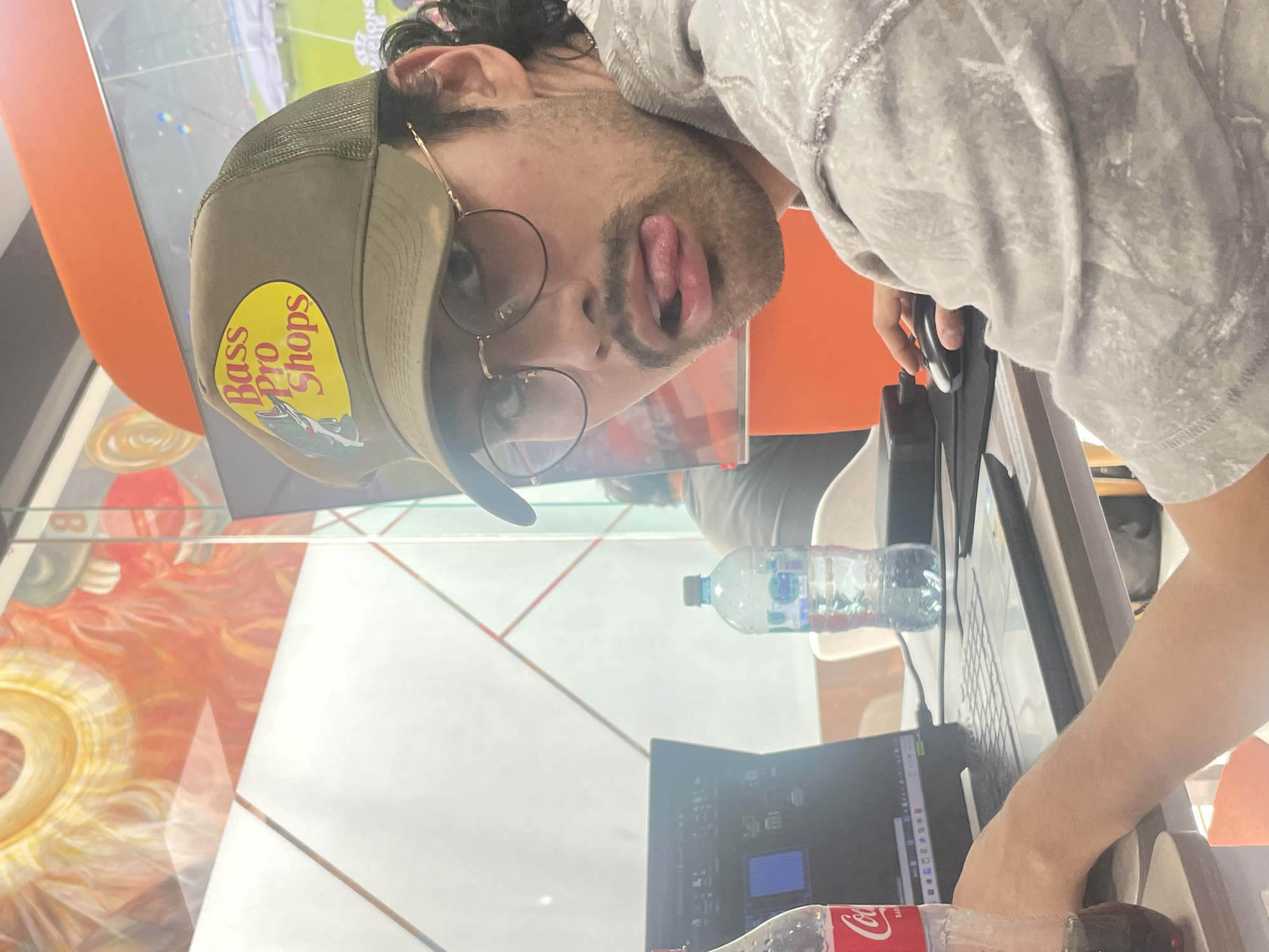 A male is indoors at a desk, likely a public space in San Andrés Cholula, Mexico, based on interior design and observed objects. He wears a gray hoodie and a Bass Pro Shops cap, round-rimmed glasses, and has a goatee. He is sticking his tongue out at the camera. He is using a computer with a wired mouse. A bottle of water and a bottle of Coca-Cola are nearby on the desk. The backdrop includes colorful artwork and an orange wall. A TV screen in the background displays a soccer game.