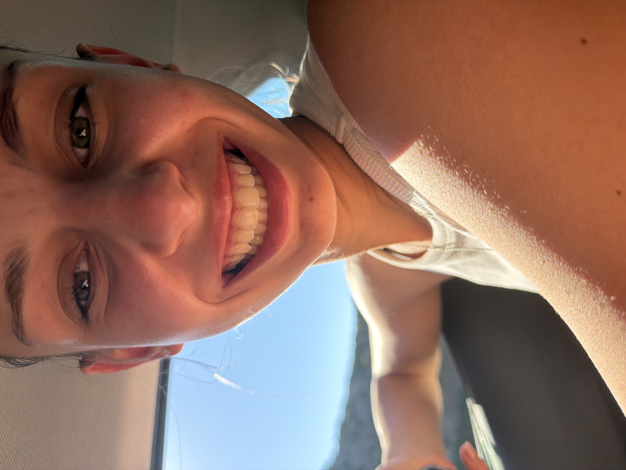 A close-up, low-angle shot shows a smiling Caucasian woman with light skin. Her eyes are brown, and her teeth are visible in a wide smile. She is wearing a white tank top. The background features a clear blue sky, suggesting an outdoor setting. The presence of a car interior (possibly a seat or panel) indicates that the photo was taken inside a vehicle. The city of Escorca, Spain, is not directly discernible from the visual information.