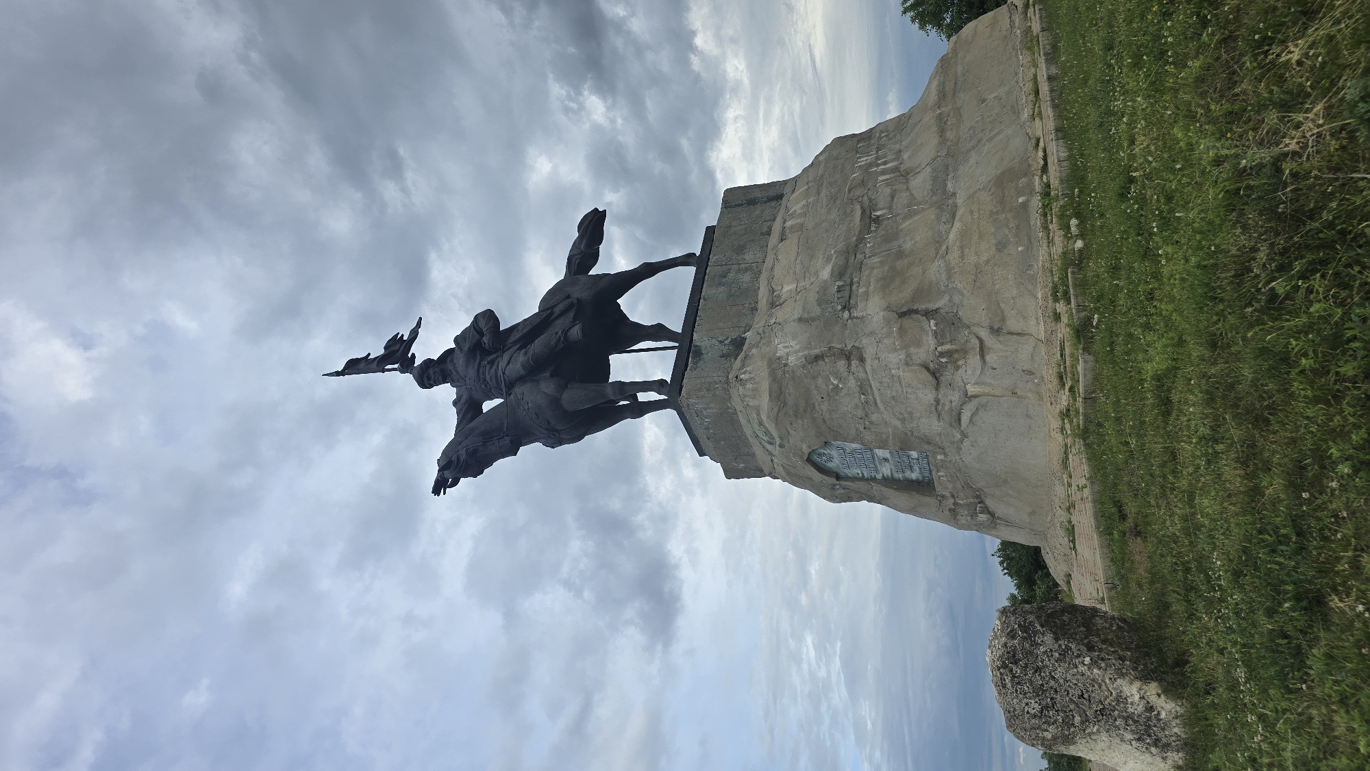 This media file shows a statue of a man on horseback, located in Yelabuga, Russia. The statue is made of dark metal and depicts the man holding a flag or banner aloft with his left hand. He is dressed in historical attire, including a hat and a coat. The horse is shown in mid-stride, with its head turned slightly.
The statue is mounted on a large, rough-hewn stone pedestal that appears to be part of a larger rock formation. The pedestal has some visible carving or inscription, though the text is not entirely legible in the image. To the right of the pedestal, there is a grassy slope dotted with small white flowers. The background is dominated by a cloudy sky, with patches of blue visible. The overall impression is that of a monument set in an outdoor, possibly historical or memorial, location. The lighting suggests it is daytime, but the heavy cloud cover might indicate an overcast or approaching storm. The orientation of the image is vertical, and the scene is viewed from a low angle, emphasizing the scale of the monument.
