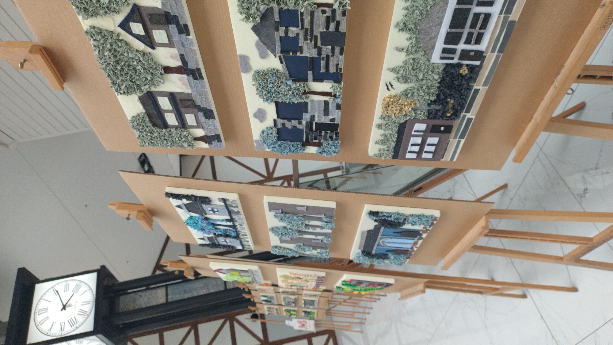 This is a medium shot, eye-level, indoor photograph of an art exhibition with multiple easels displaying textured artwork. The background features a large clock tower and a ceiling with a geometric pattern. The art pieces are displayed on brown cardboard panels, each featuring textured representations of houses and trees. The artwork appears to be made of various materials creating a relief effect. The floor is a tiled marble pattern. The image does not contain any people. The location is identified as the city of Jelgava, Latvia, though no specific landmarks other than the clock tower are visible to confirm this.