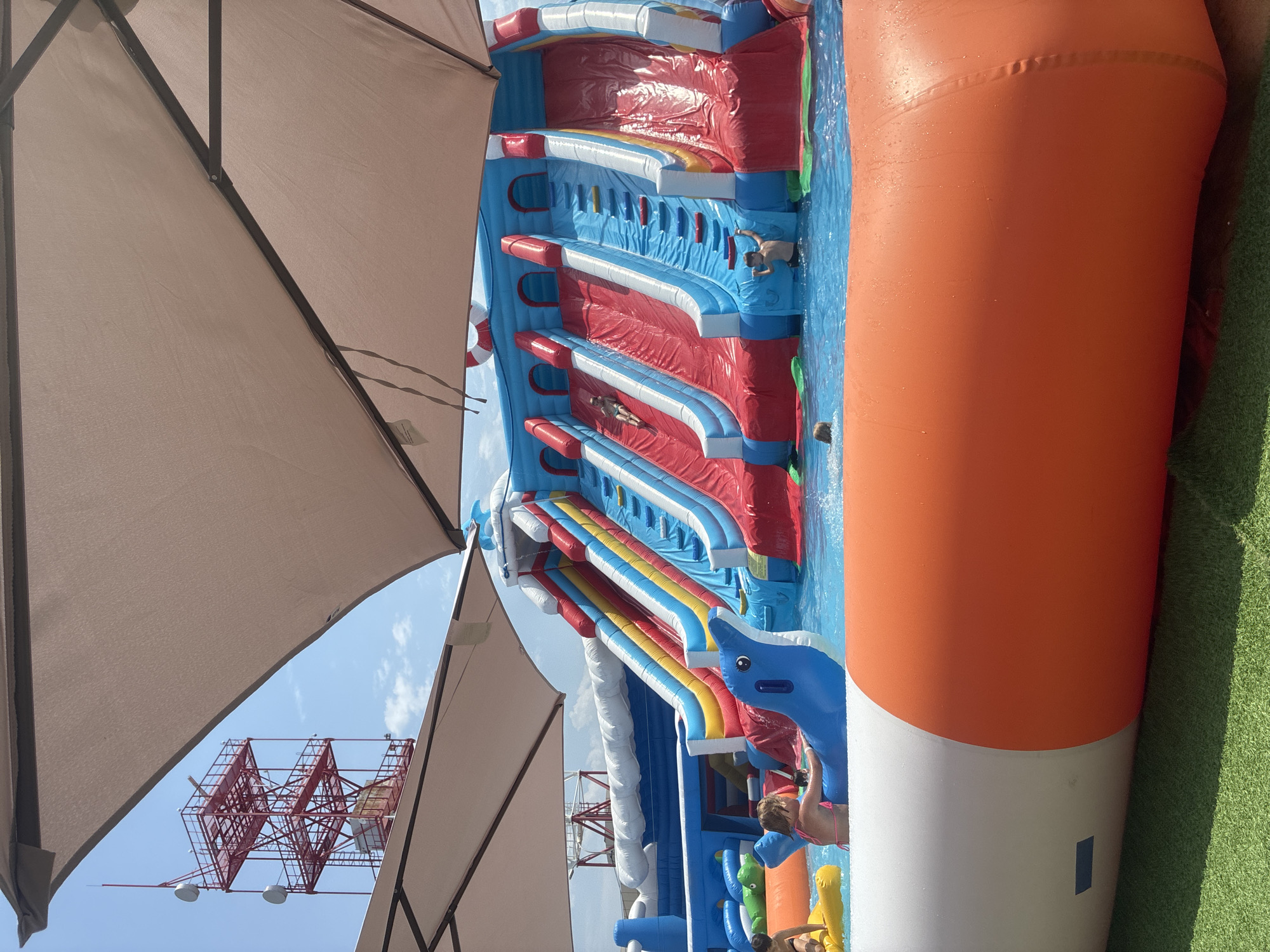 An aerial inflatable water park is set up on a sunny day, with several children interacting with the slides and pool. The park features multiple slides with red, blue, yellow, and white sections, a blue inflatable dolphin, and an orange and white inflatable bumper. In the background, a large red transmission tower is visible against a blue sky with scattered clouds, suggesting an outdoor location. The area is covered with green artificial turf in the foreground.