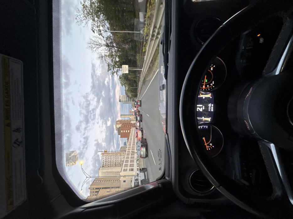 The image provides a driver's perspective from inside a vehicle, likely a Jeep Wrangler, on a multi-lane highway in Boston, Massachusetts, United States. The vehicle is traveling southbound on I-93 (Southeast Expressway), with the view extending towards the South End and the broader Boston skyline.
The time of day is late afternoon or early evening, indicated by the overcast sky with scattered clouds and diffused natural light. The vehicle's instrument cluster shows a speed of 21 mph, the transmission in 'PRND', and an odometer reading of 23,191 miles. The fuel gauge indicates approximately one-quarter of a tank.
Ahead, the highway carries moderate traffic, with several passenger vehicles, including a white Jeep Wrangler in the adjacent left lane, moving at a slow speed. Brake lights are illuminated on some vehicles in the distance.
On the left side of the highway, a concrete barrier separates the roadway from urban development. Visible structures include a large, light-colored building with numerous windows, followed by a cluster of red-brick buildings, and a modern high-rise tower further in the distance, contributing to the city skyline. A streetlight is visible above the left lanes.
On the right side of the highway, another concrete barrier runs parallel to the road, separating it from a lower-lying area. This area is densely populated with mature trees and foliage. A white rectangular sign with black text reading "EMERGENCY STOPPING ONLY" is mounted on a pole within this treed section. Partially visible through the trees are elements consistent with a railway line, including tracks and signaling equipment.
The upper portion of the vehicle's windshield displays a faded, rectangular adhesive residue or sticker, and the overhead visor area contains a "WARNING / MISE EN GARDE" label.