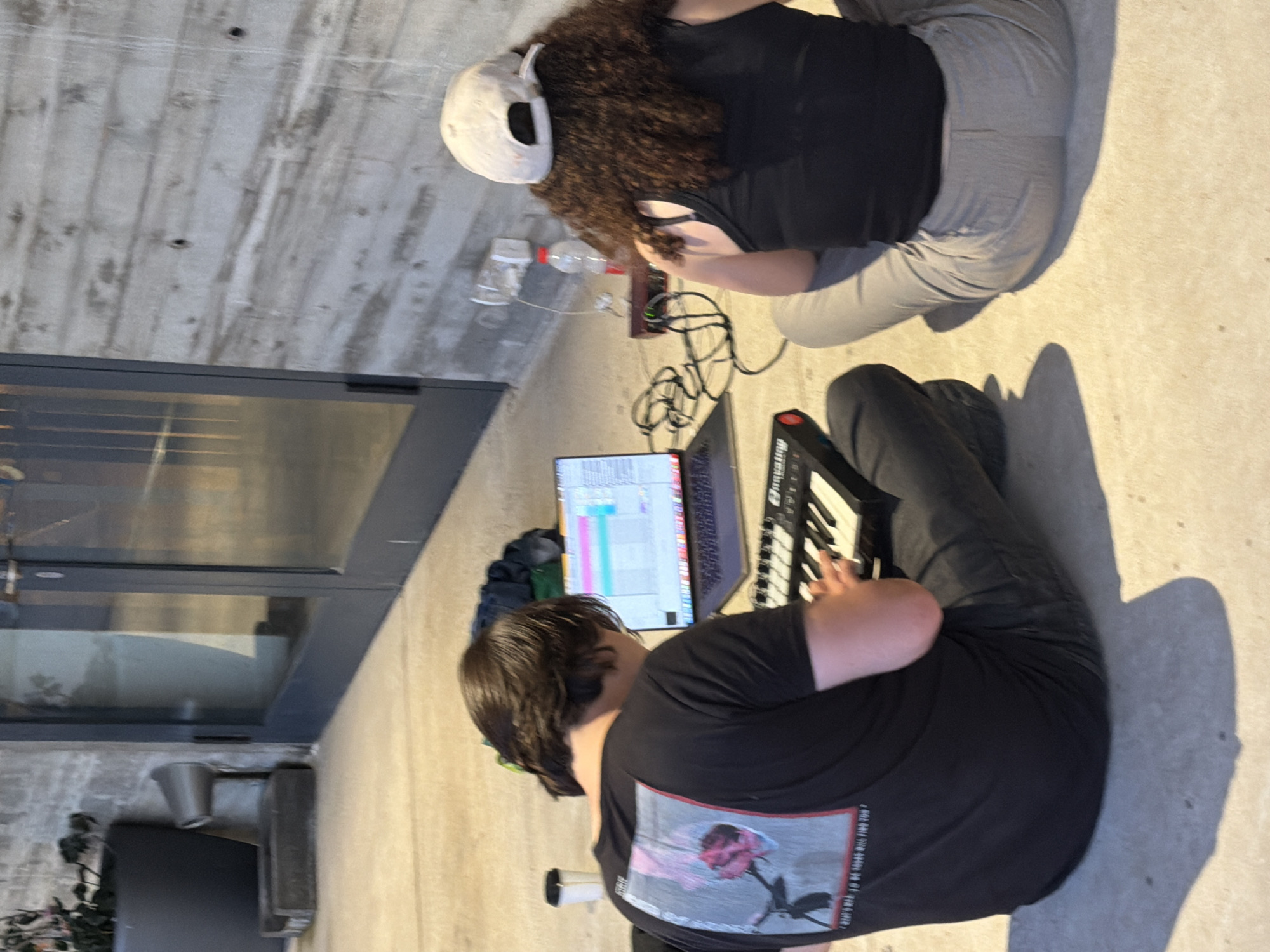 The image captures two individuals engaged in music production in an outdoor setting. One person, wearing a white cap, is seated with their back to the camera. They have curly brown hair and are wearing a black shirt and light gray pants. Beside them are water bottles and connected electronic devices. The second person, facing away from the camera, is seated and playing a small electronic keyboard connected to a laptop. This individual has short brown hair and is wearing a black t-shirt with a rose graphic on the back and dark pants. The setting is an outdoor patio or deck, and possibly in Tel Aviv, Israel. There is a door or window in the background, suggesting the location is part of a building. The lighting suggests it is daytime.