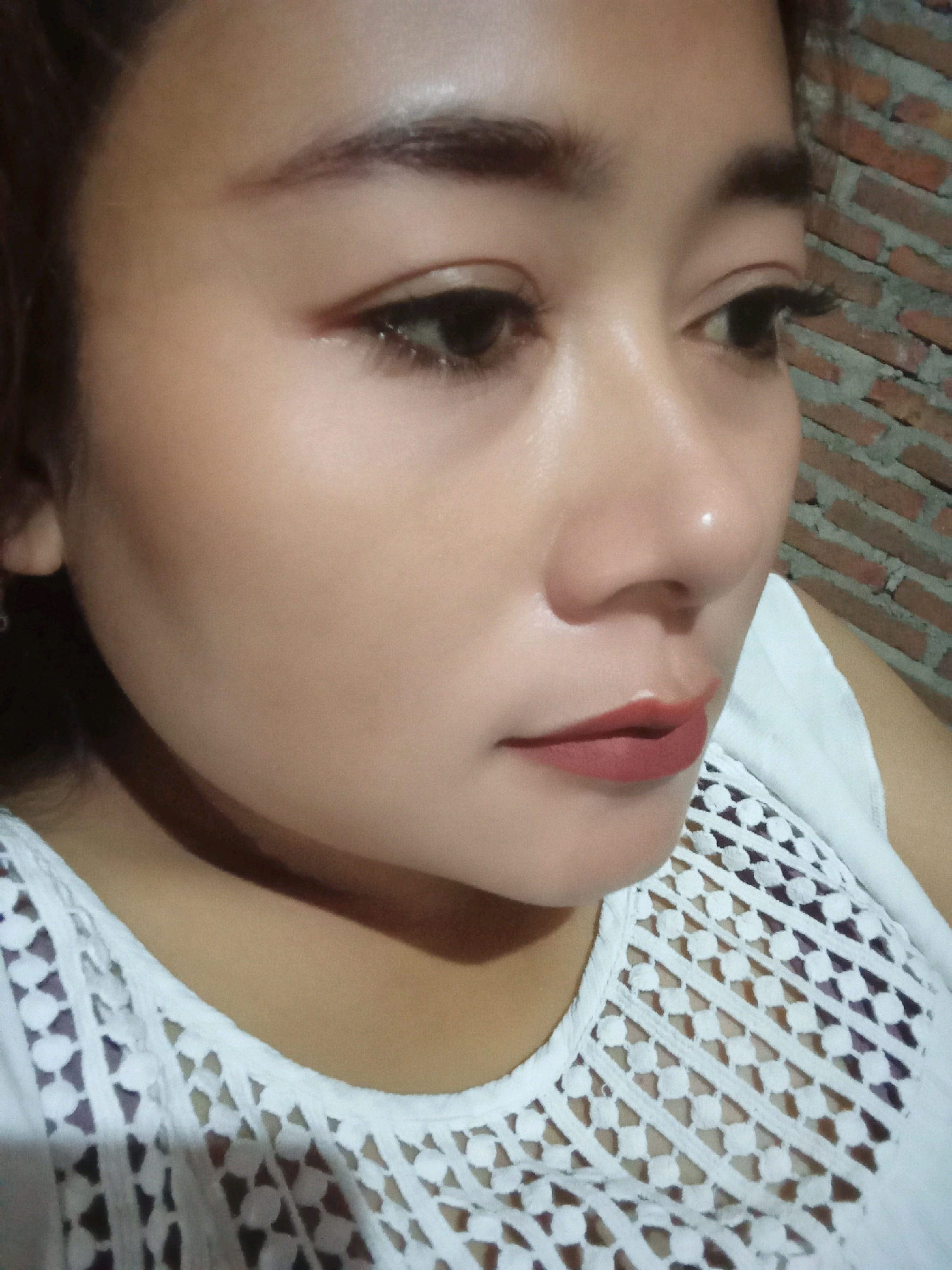 The image shows a close-up of a young woman's face, tilted to her right. She has dark hair pulled back, revealing her features. Her makeup includes foundation, subtle eyeshadow, defined eyebrows, and matte lipstick in a muted red shade. She is wearing a white, sleeveless top with an intricate lace or crochet design around the neckline. The background consists of a brick wall, suggesting an indoor or semi-outdoor setting. The lighting appears to be artificial, casting a slight sheen on her skin. The image has a grainy quality.