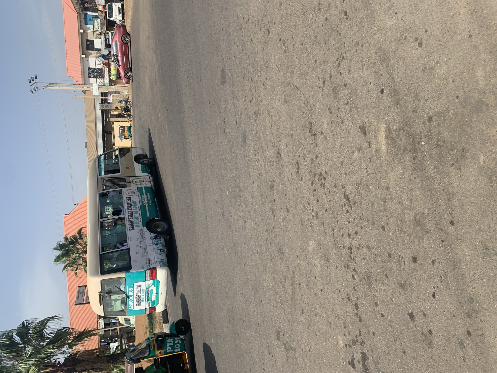 The image depicts a street scene in Rafin Par, Nigeria. A "BRIGHTSTARS EXCELLENT HEIGHT ACADEMY" school bus is parked on the side of the road, along with a three-wheeled vehicle labeled "PTA 1037". Buildings, power lines, palm trees, and other vehicles are visible in the background.