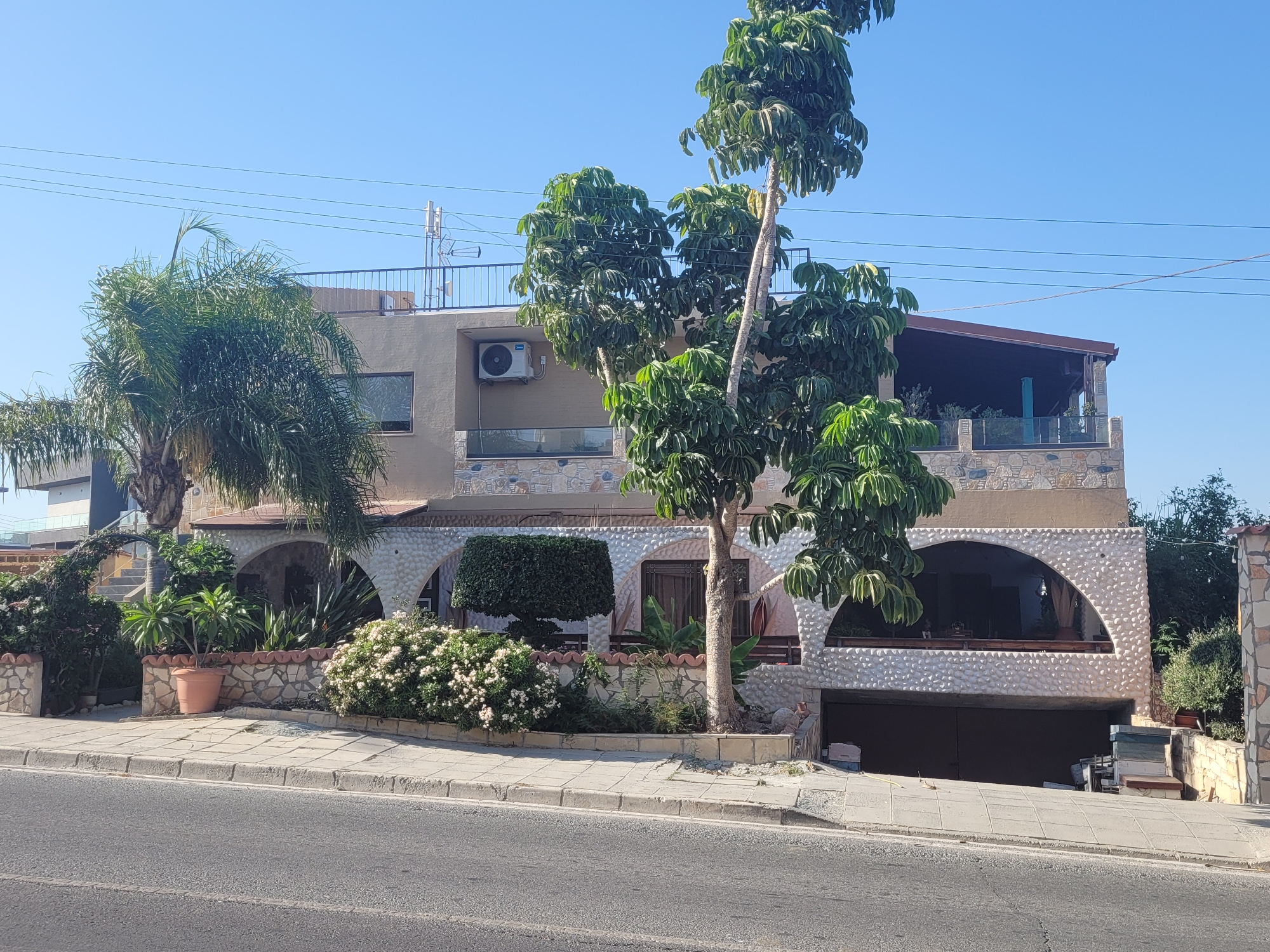 A two-story residential building with a tan-colored facade and a brown tiled roof is situated on a street in Episkopi, Cyprus. The building features several arched openings, some of which are covered with light-colored, textured material, and others with windows or balconies. A large, dark garage door is visible at the ground level. The property is adorned with various plants, including palm trees, flowering bushes, and a neatly trimmed hedge. A paved sidewalk runs along the front of the building, separating it from the asphalt road. Overhead power lines are visible against a clear blue sky. The scene appears to be captured during daylight hours.
