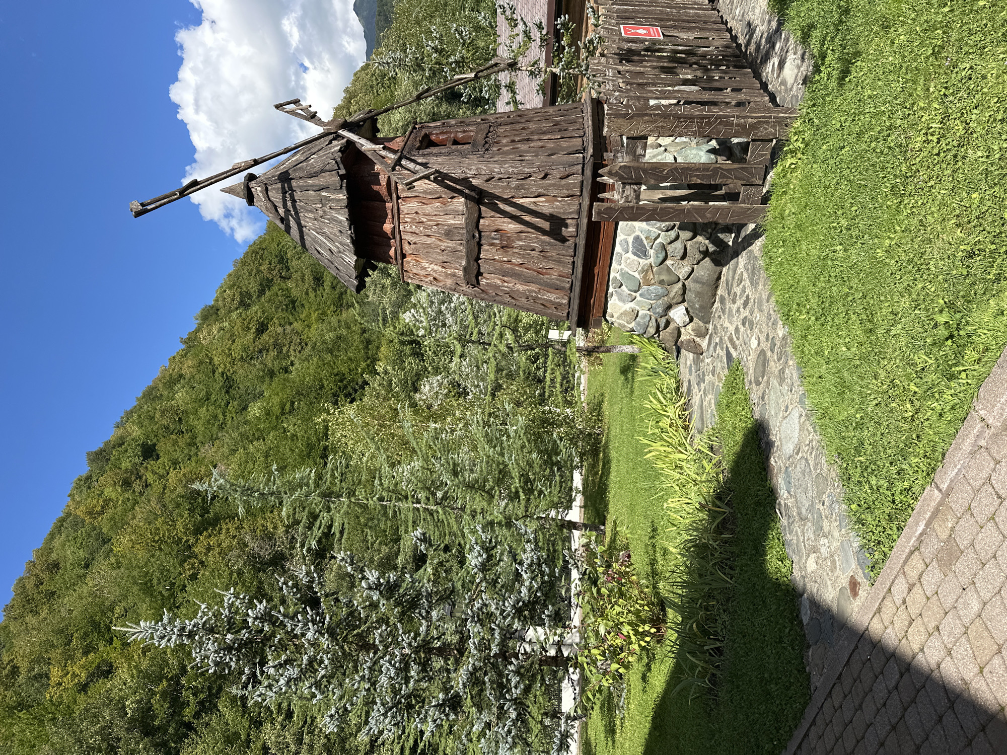 This is a high-angle, full-shot photograph taken in Estosadok, Russia. The scene depicts a rustic, wooden windmill situated in a lush green garden with a mountain covered in dense forest in the background. The windmill is crafted from weathered wood, with a conical roof and two prominent blades, one of which is broken. Its base is constructed from natural stones. A rustic wooden fence surrounds the windmill, and a small red sign with a white silhouette is attached to it. The foreground shows a paved walkway made of beige rectangular stones, transitioning to a stone pathway leading to the windmill. The garden is neatly manicured with vibrant green grass and various plants and trees, including tall evergreen trees. The sky is a clear blue with a few scattered white clouds. The overall impression is of a serene, natural setting with a charming, handcrafted focal point.
