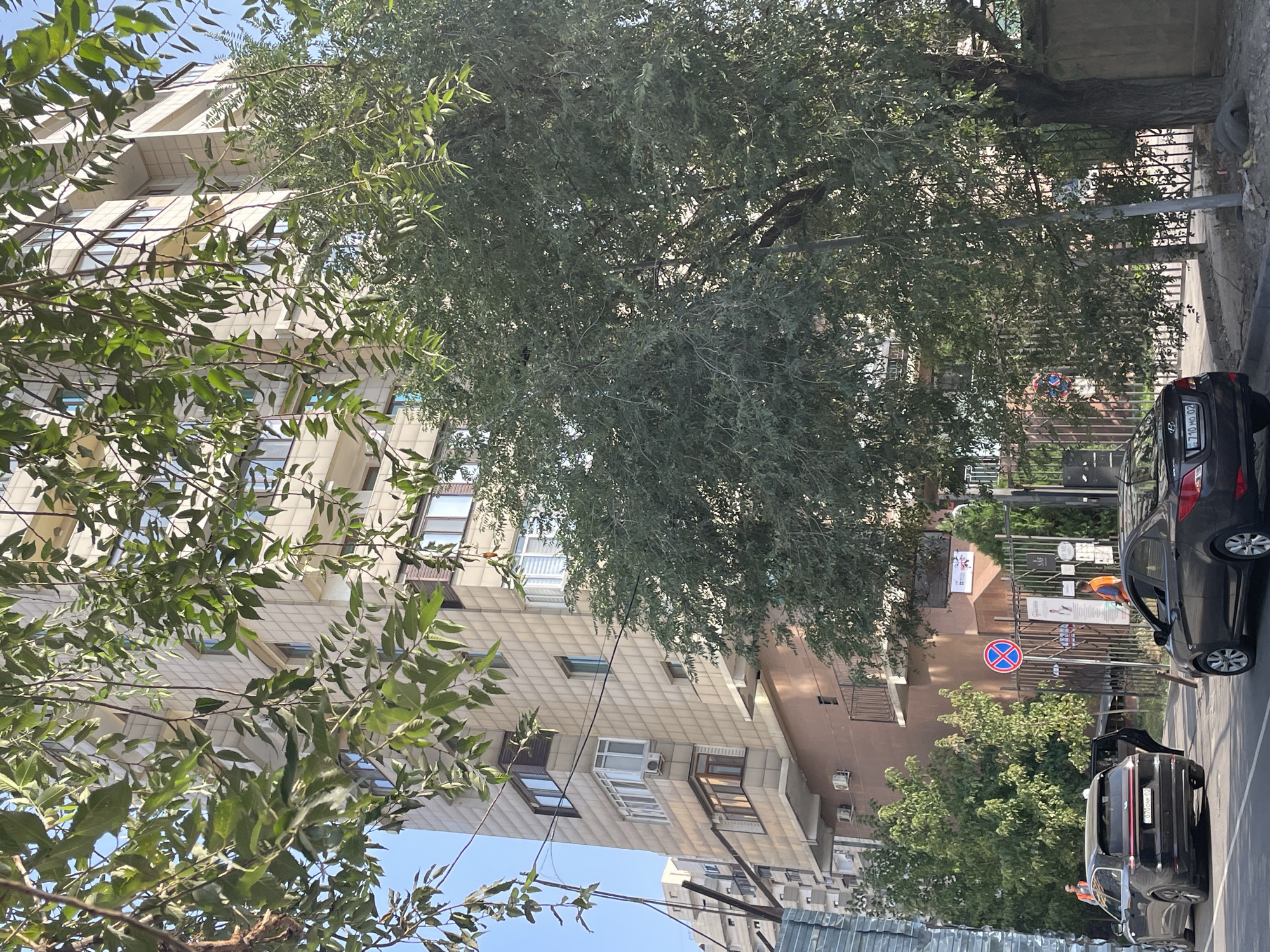 The image depicts a street scene in Almaty, Kazakhstan, likely in a residential or mixed-use area. A tall, modern apartment building with a light-colored, tiled facade dominates the background. Trees with lush green foliage partially obscure the building, suggesting a well-treed urban environment. In the foreground, two cars are parked on the street: a black SUV with its rear door open and a dark sedan. Several individuals are visible near the cars and the entrance to a property, some wearing bright orange vests, possibly indicating they are workers or security personnel. A traffic sign with a red circle and a diagonal cross is present, along with a fence and the entrance to a building. The overall impression is of a sunny day in an active urban setting.