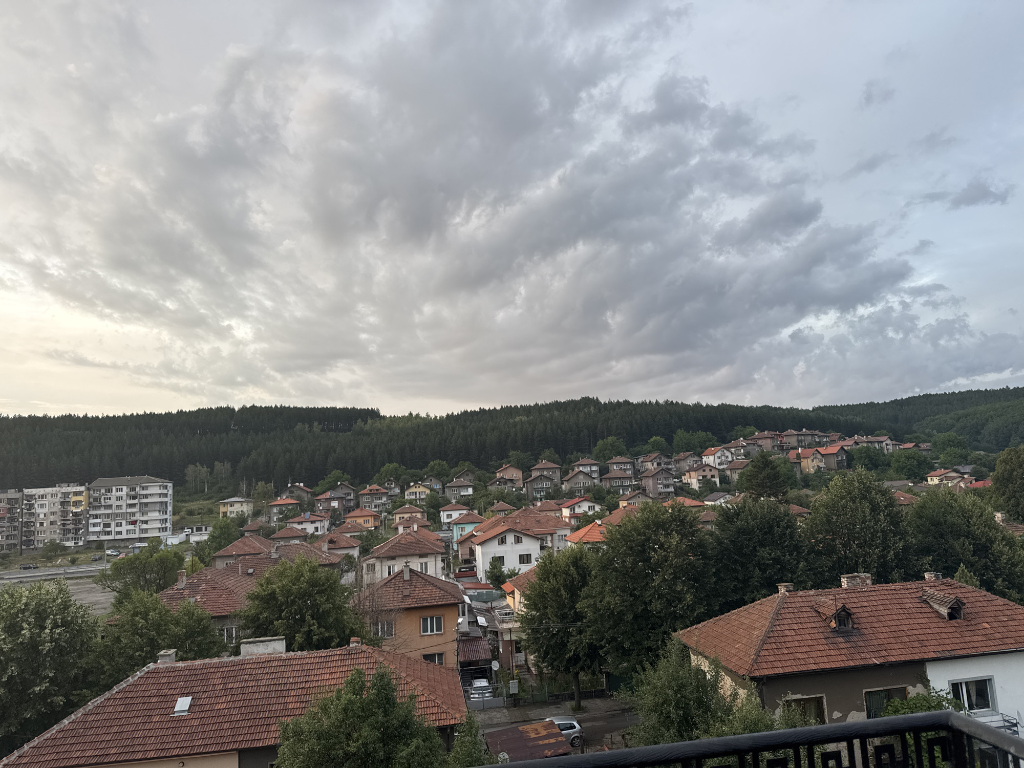 This is a high-angle shot of a residential area in Pernik, Bulgaria. The sky is overcast with gray clouds. The foreground is dominated by the red-tiled roofs of houses, some with dormer windows and chimneys. Mature green trees are interspersed among the buildings.
In the mid-ground, more houses with red-tiled roofs are visible, built on a hillside. A multi-story apartment building, gray and white with numerous windows, stands on the left side of the frame, overlooking a road. Several cars are parked on the road and in a lot adjacent to the apartment building.
The background features a dense forest of dark green pine trees covering a hill. The overall impression is of a somewhat aged, densely populated suburban or town environment nestled within a hilly, forested landscape. There are no people visible in the scene.