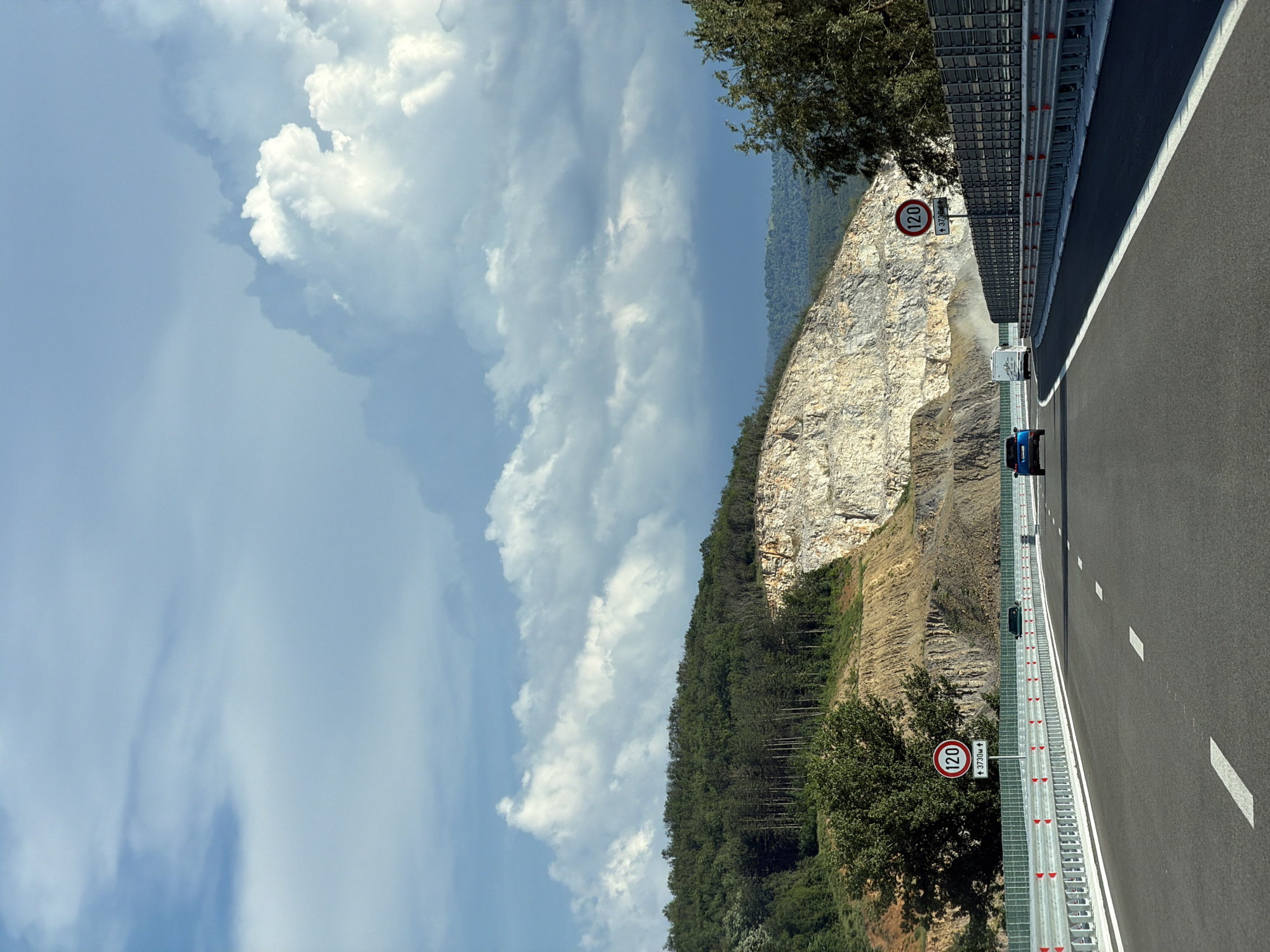 The image depicts a highway in a mountainous region, with a sheer rock face on one side and a steep, wooded slope on the other. The highway is paved with asphalt and marked with white lane dividers. A metal guardrail runs along the side of the road, equipped with reflective markers.
On the highway, a white van is visible in the distance, followed by a blue car. A speed limit sign indicating "120" is prominently displayed next to the road. Another similar sign is visible further down the road. The sky is partly cloudy, with large, white cumulus clouds against a pale blue background. The time of day appears to be daytime, likely late morning or early afternoon, given the lighting. The overall scene suggests a scenic drive through a natural landscape, possibly a quarry or a mountainous pass.
The location context provided is Peshterna, Bulgaria. The prominent rock face suggests a geological feature that might be a natural cliff or a result of quarrying activities. The dense pine forest on the slope indicates a temperate climate. The presence of the highway suggests a connection to human infrastructure facilitating travel through this terrain. The weather appears pleasant, with good visibility.