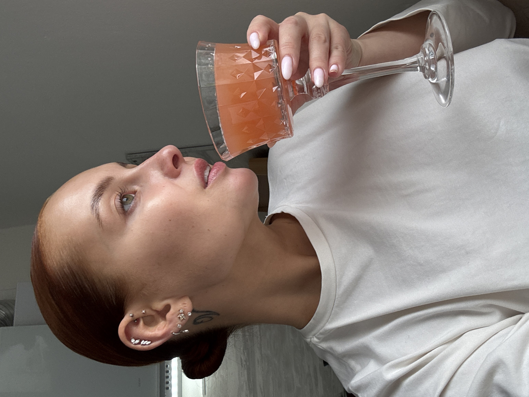 A fair-skinned woman with auburn hair pulled back into a low bun is holding a clear glass with an orange-pink liquid, appearing to drink from it. She is wearing a white long-sleeved shirt. Her ear is pierced in multiple locations, and a tattoo is visible behind her ear. The background is muted, showing an off-white wall and what appears to be a refrigerator. There is no contextual information in the image indicating that it was taken in the city of Chaikovsky, Russia.