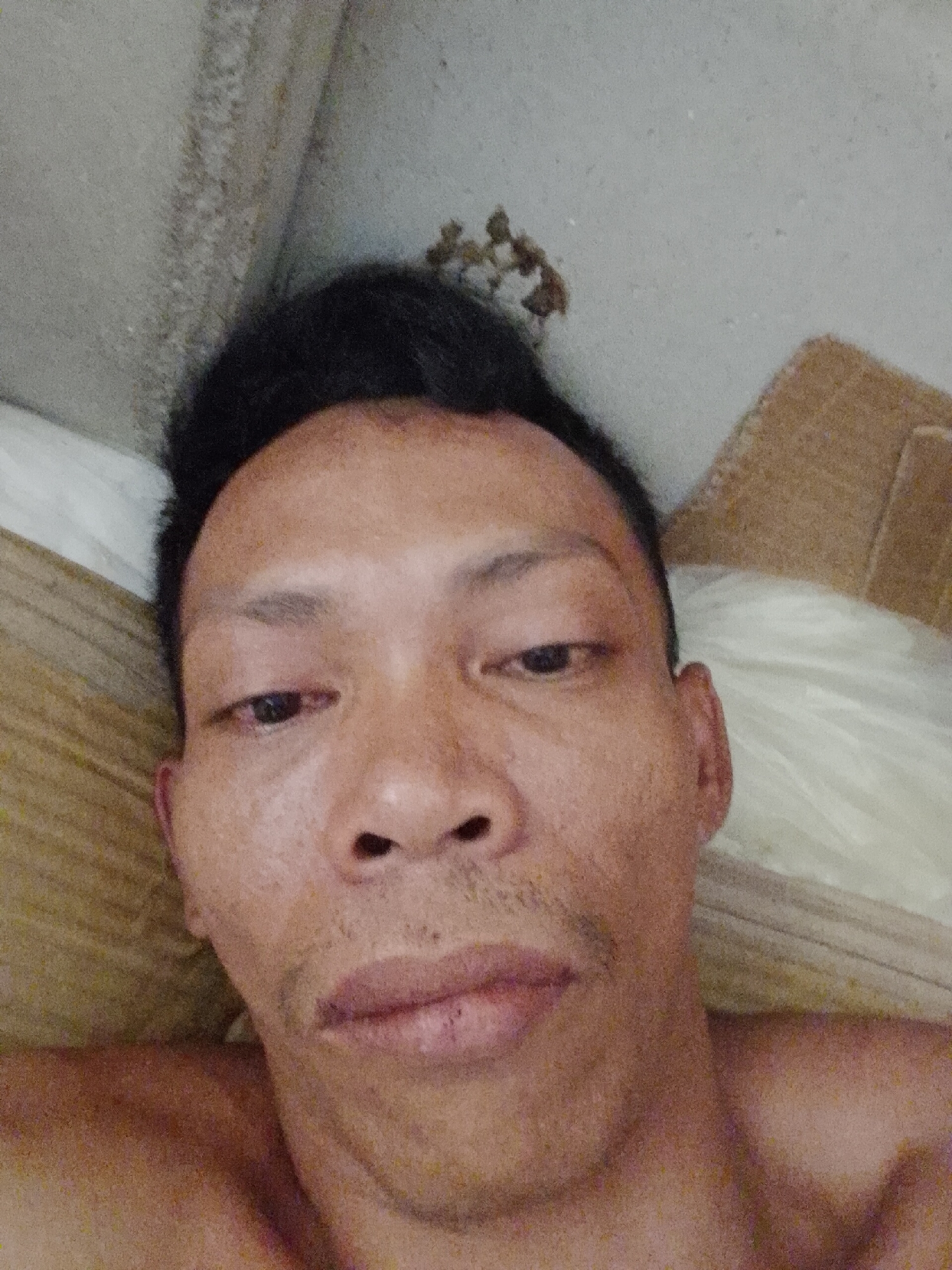 A close-up, selfie-style image depicts a man with dark hair lying down. The man has brown skin, short, dark hair styled upwards, and stubble on his upper lip and chin. His eyes are open but appear tired. He is shirtless. The background is indistinct but shows what appears to be a wall and some bedding. There is some unknown debris on the wall near the man's hair. The location is noted as Bukit Ketok, Indonesia.
