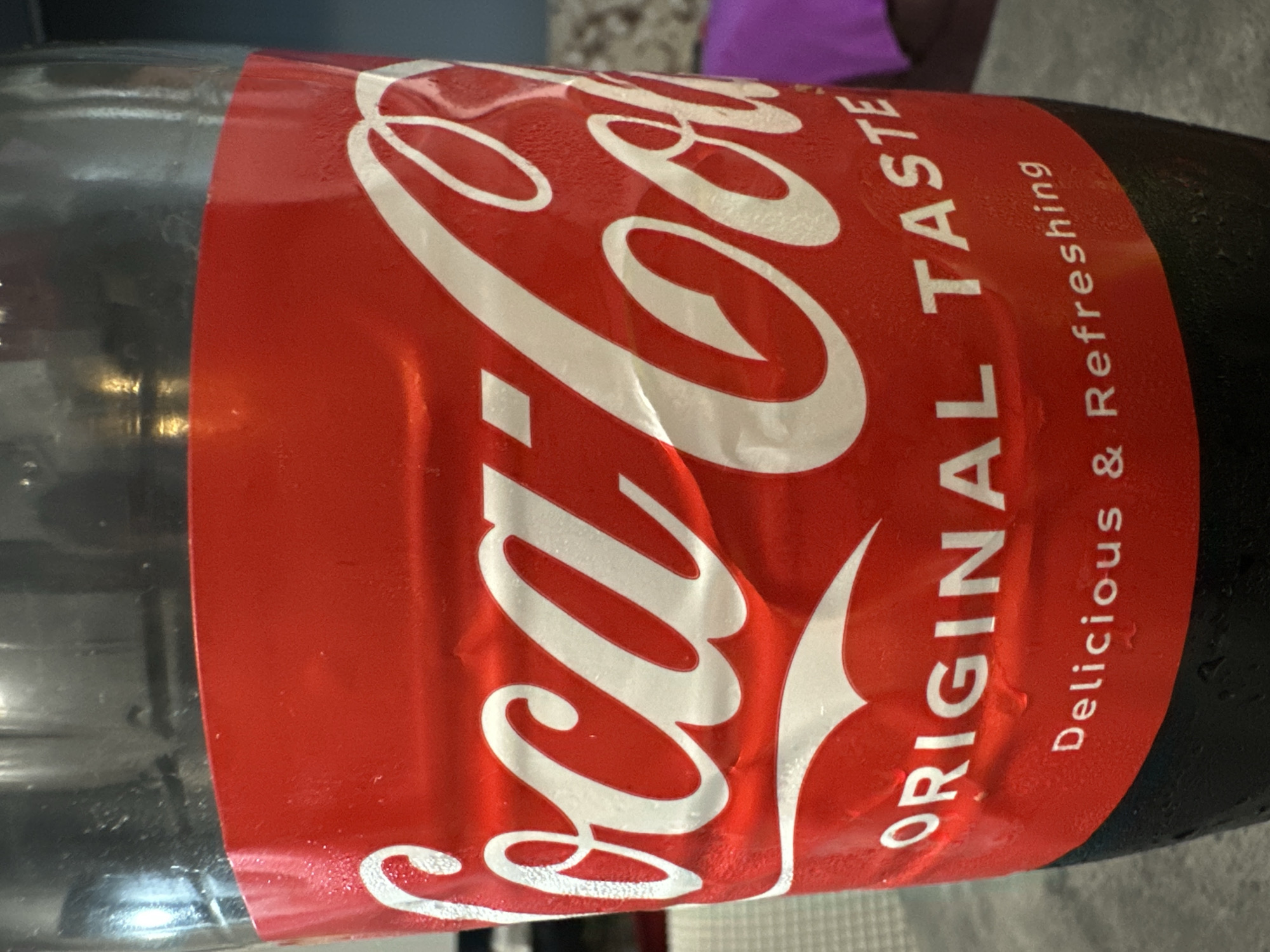 A close-up, eye-level shot shows the label of a Coca-Cola bottle. The label is bright red and features the iconic white Coca-Cola logo in large script. Below the logo, "ORIGINAL TASTE" is written in white, followed by "Delicious & Refreshing" in a smaller font. The bottle appears to be sweating, with condensation visible on the surface. The background is blurred, but suggests an indoor setting with a gray countertop in the foreground and hints of other objects and a person in purple clothing in the background. The image does not provide any specific visual cues to indicate a location in Varna, Bulgaria.