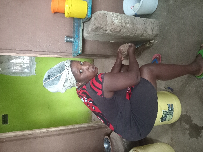 A woman is depicted in an indoor or semi-enclosed utility space, likely a kitchen or work area, in Manda-Shivanga ward, Kenya. She has dark skin and is wearing a dark, sleeveless top with red patterns, dark shorts, and a light-colored head covering. She is shown in a squatting or seated position on an upturned yellow plastic bucket, looking directly at the camera with a neutral expression.
With both hands, she is holding a small, rough, irregularly shaped object, possibly a coconut, appearing to be in the process of grating or preparing it. Near her bare feet, red and green flip-flops are visible on the concrete floor. A silver bowl and another yellow container are also on the floor nearby.
The walls are painted in two tones: an upper brown/beige and a lower vibrant green. A window on the green wall provides natural light, indicating it is daytime. Above the window line, on a raised counter-like structure made of concrete blocks and a wooden plank, are orange and yellow plastic buckets and a metallic pipe or spigot. A small broom rests on the floor near this structure. The floor is concrete. Partially visible text on the yellow bucket she is sitting on includes "GIANT GR..." and "PROD...".