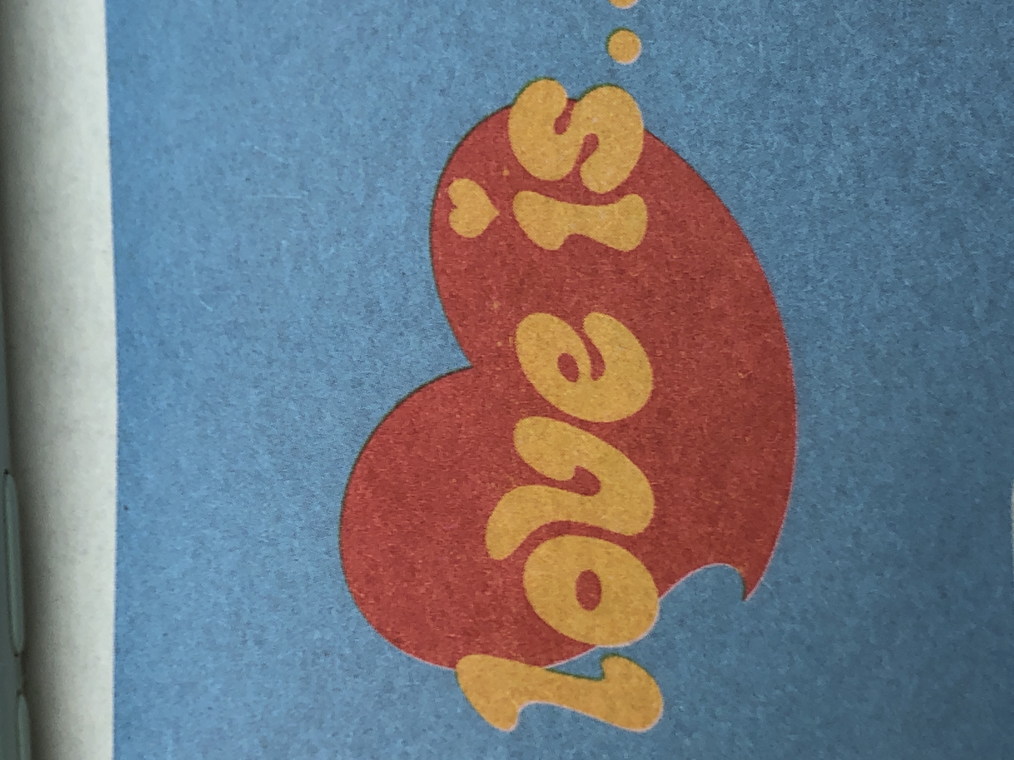 The image presents a close-up view of a vibrant, textured graphic design. The background is a muted, speckled blue, suggesting a fabric or textured paper surface. Centered prominently within the frame is a large, stylized heart shape in a deep red hue.
Inside the red heart, text in a bright yellow-orange color is arranged vertically. It reads "Love is," with "Love" on the left side and "is" on the right. A notable detail is that the dot above the 'i' in "is" is replaced by a tiny yellow-orange heart symbol. Above the word "is," several small, ascending yellow-orange circles, resembling bubbles, are visible. The bottom right edge of the red heart features a small, stylized indentation or "bite mark."
The entire design, including the blue background, red heart, and yellow-orange text, exhibits a slightly grainy or felt-like texture. On the far left side of the image, a narrow vertical band of light grey or white material is visible, suggesting the edge of a page or a border. There are no people or discernible activities taking place; the image is a static depiction of graphic art. No specific location cues beyond the object itself are visible.