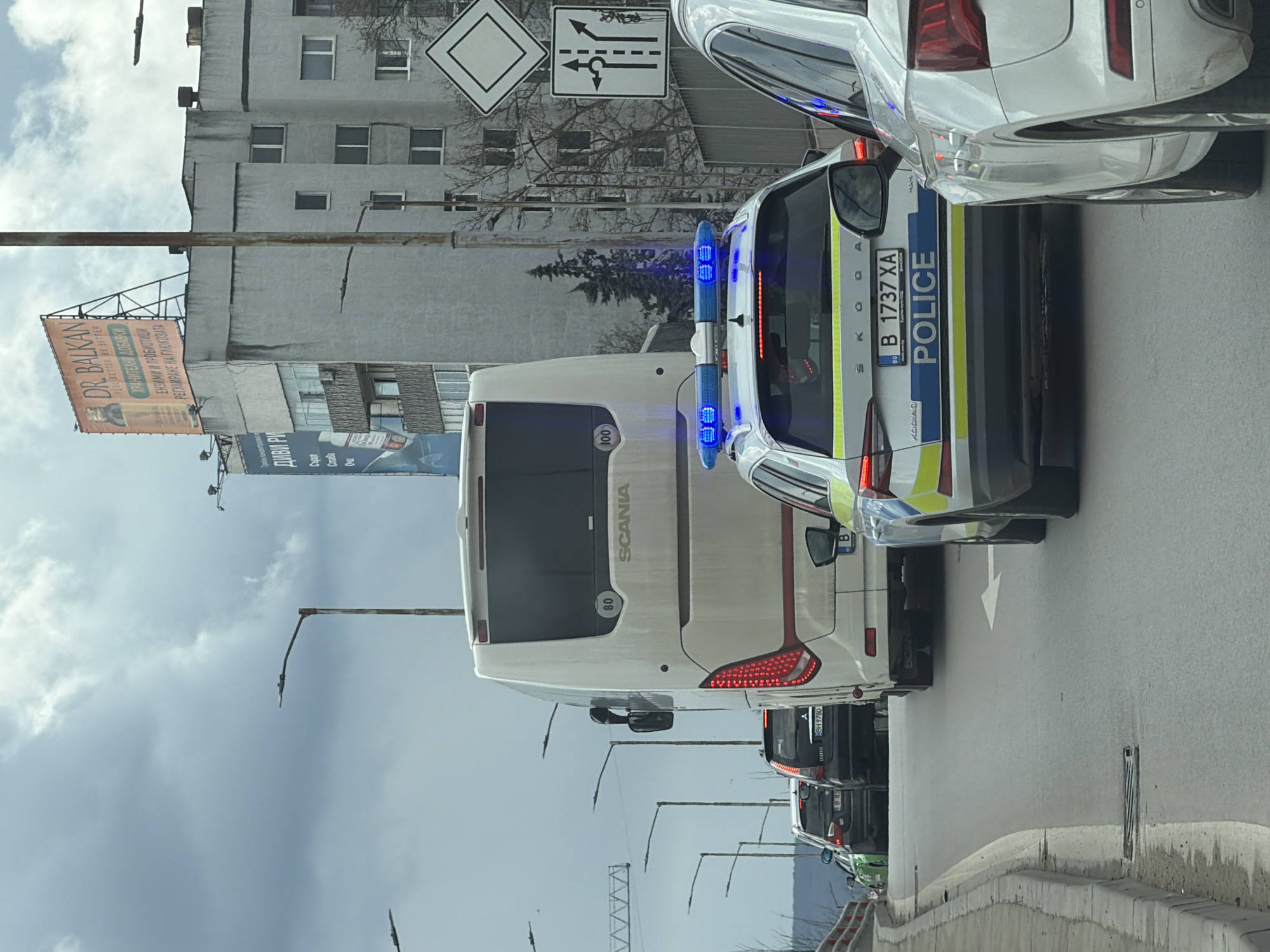 The image shows a street scene in an urban environment, likely taken in Varna, Bulgaria. A white Scania bus is in traffic, followed by a Bulgarian police Skoda Kodiaq SUV with license plate B 1737 XA. Behind the bus are other vehicles, including a black car with the license plate BH 9780 and another marked "B 83". The background includes a large billboard advertising "DR. BALKAN" and "ДИВИ РИ", a multi-story building, bare trees, traffic signs, and streetlights. The sky is cloudy.